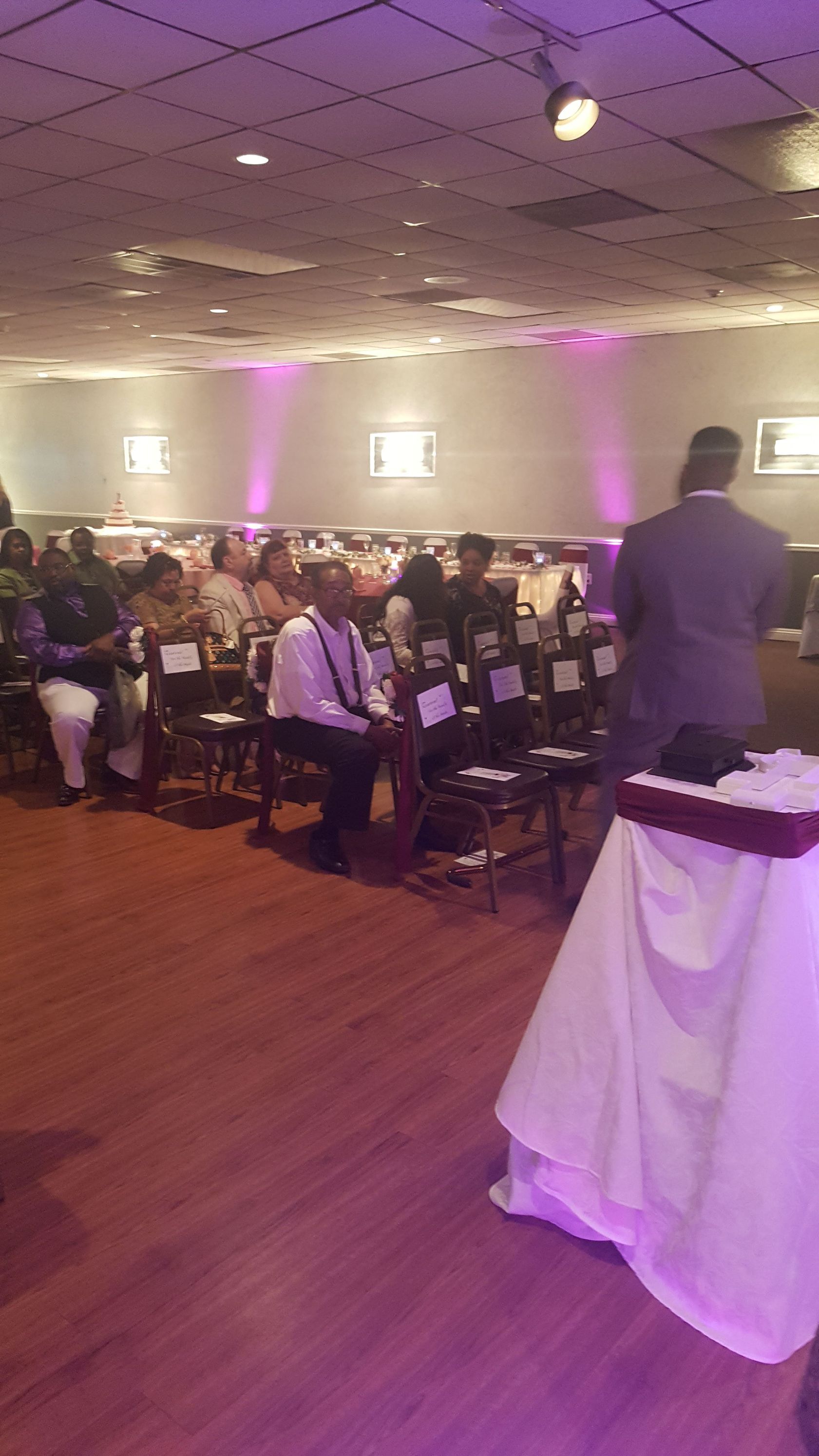 Banquet hall with seated audience facing a speaker at a podium, purple uplighting, and round tables.