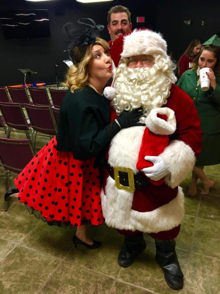 Woman in red polka-dot skirt posing with Santa at an indoor event; man stands behind them.