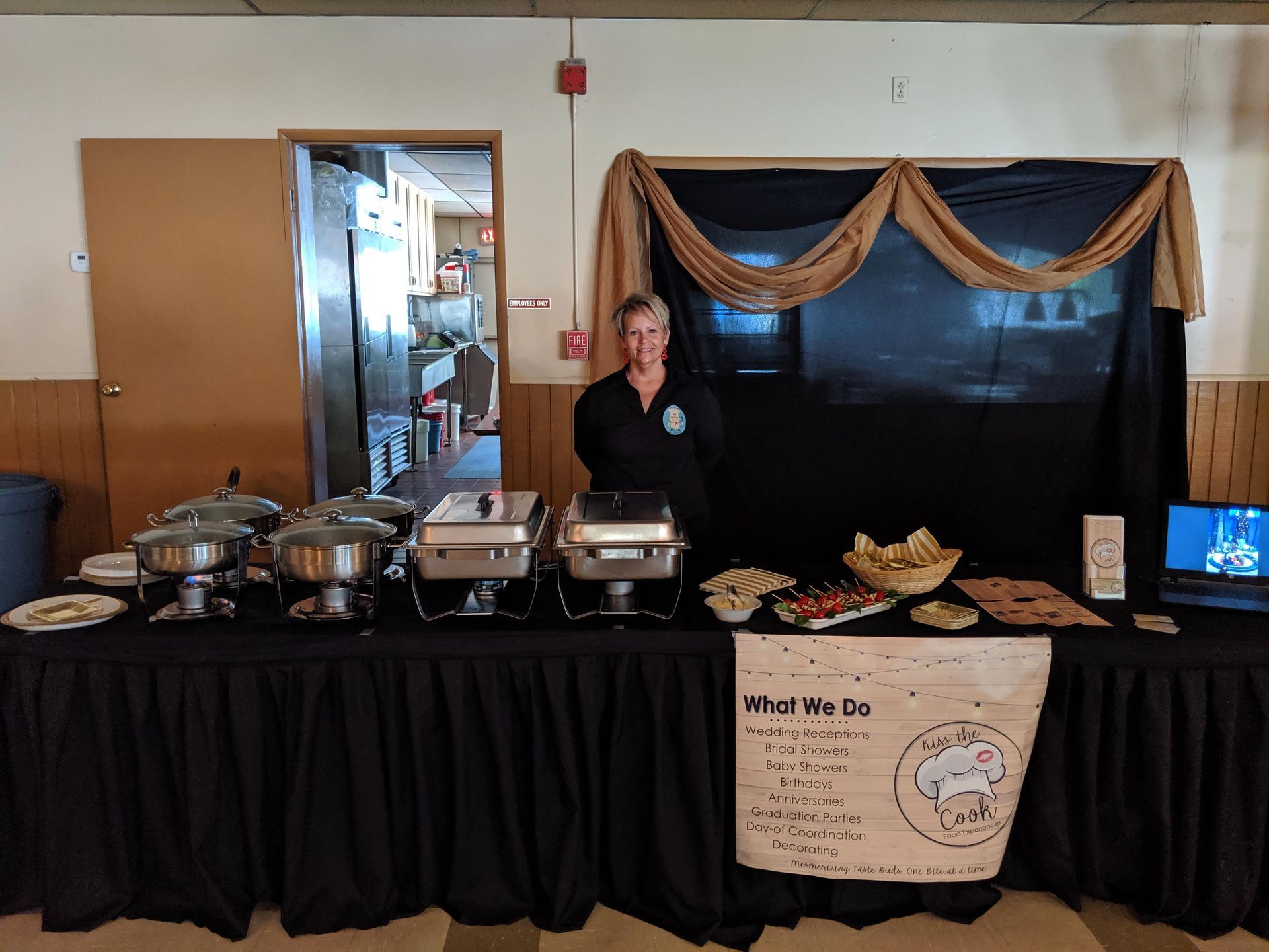 Buffet table with covered trays and a sign, with a woman standing behind it in a community room.
