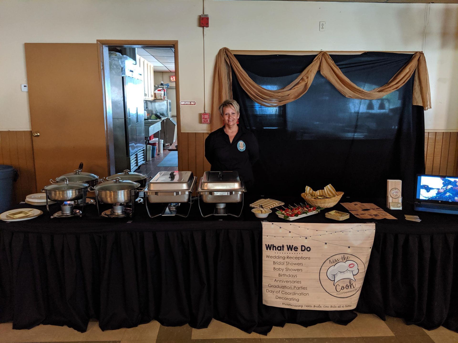Person behind a buffet table with trays of food and a “What We Do” sign at an indoor event