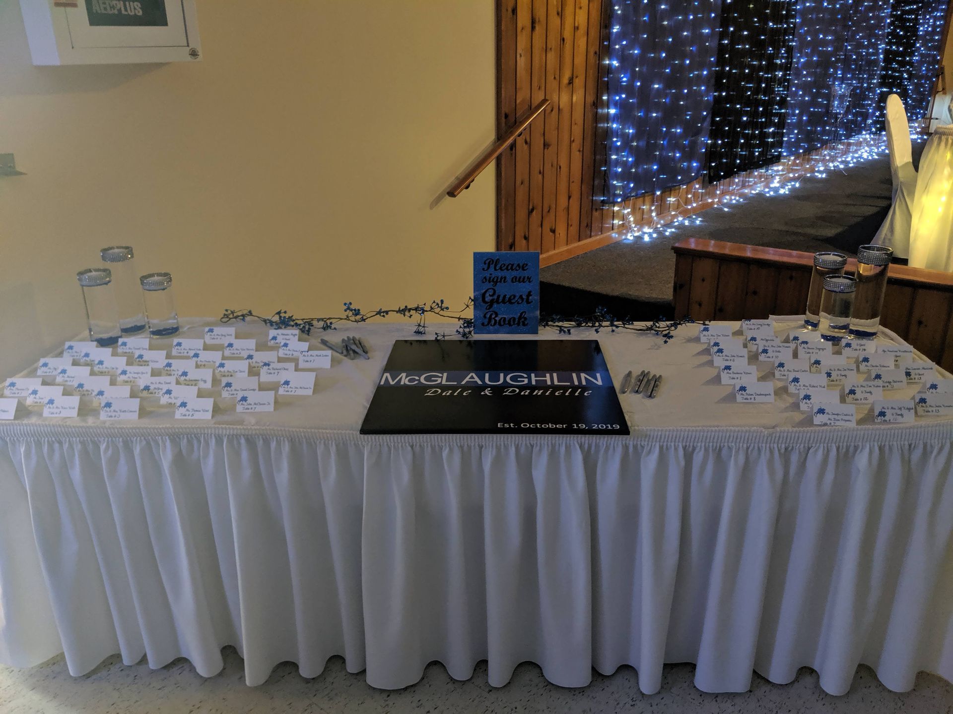 Reception table with many place cards and a black sign, draped in white at an event venue with string lights.