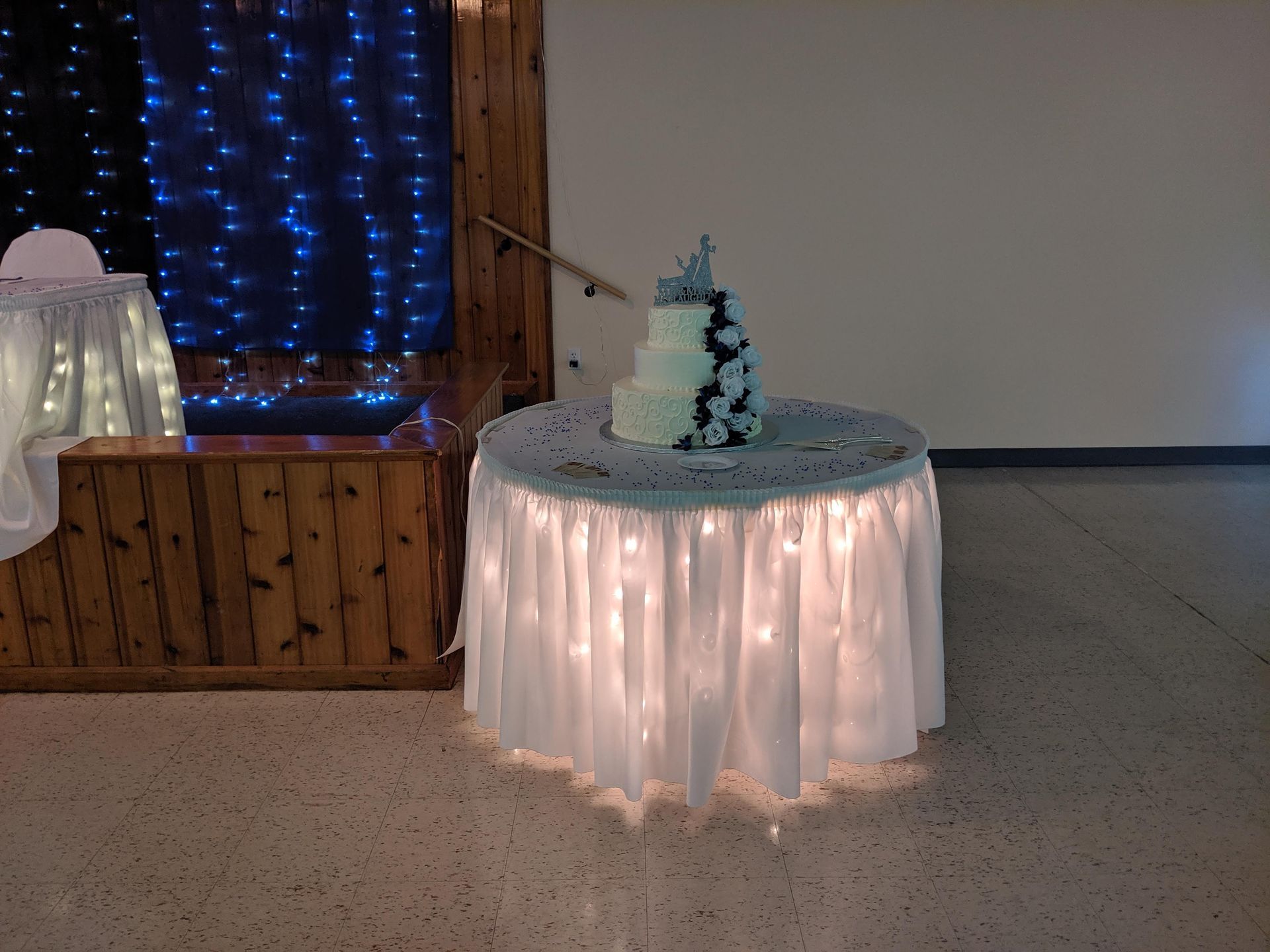 Round white-lit table with a small blue tree centerpiece in a decorated indoor venue