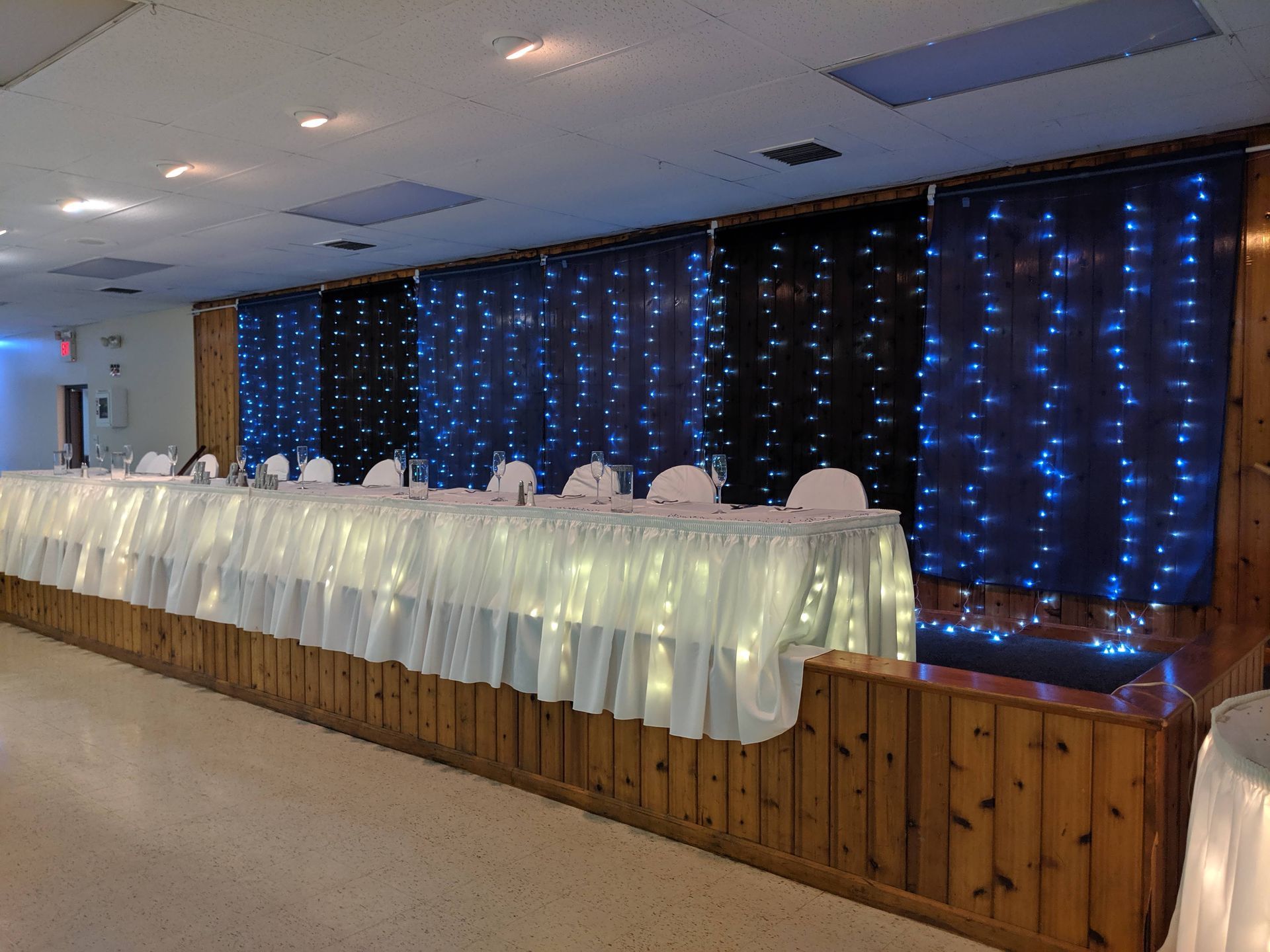 Long banquet table draped in white with blue lights and curtains in a decorated hall