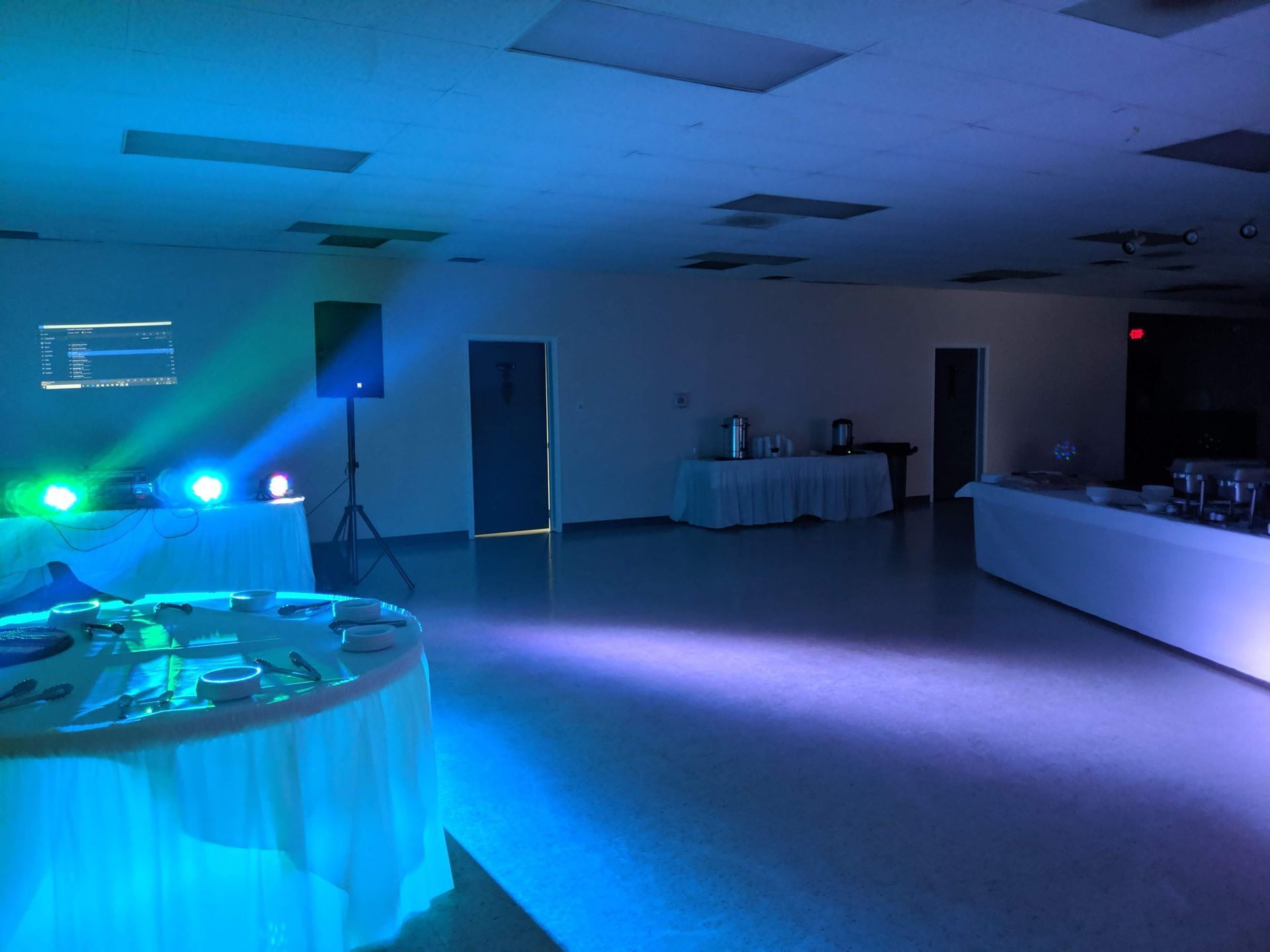 Dimly lit event room with blue stage lights, projection screen, and banquet tables at the back