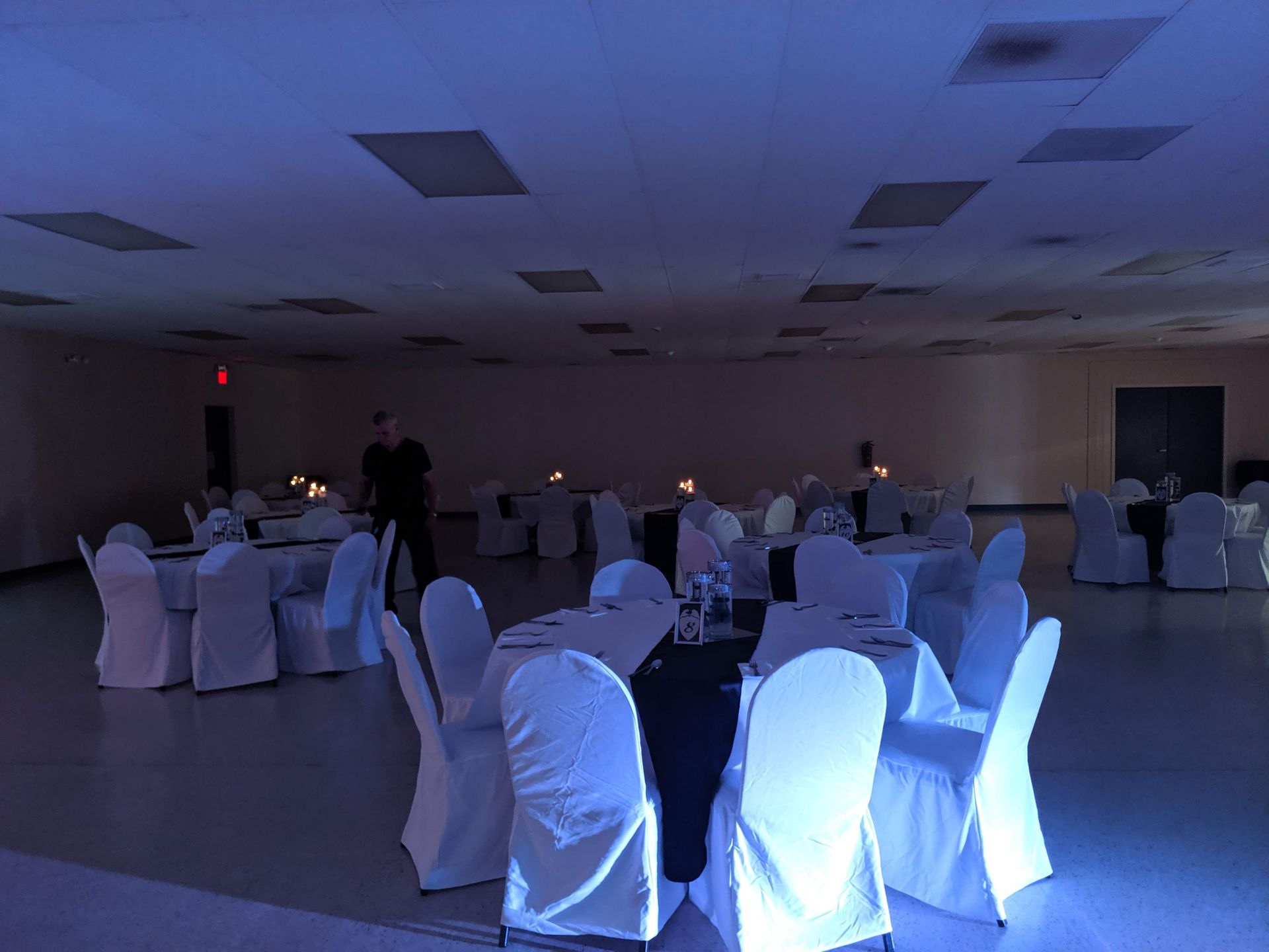 Dimly lit banquet hall with white-covered chairs around round tables and a few people setting up