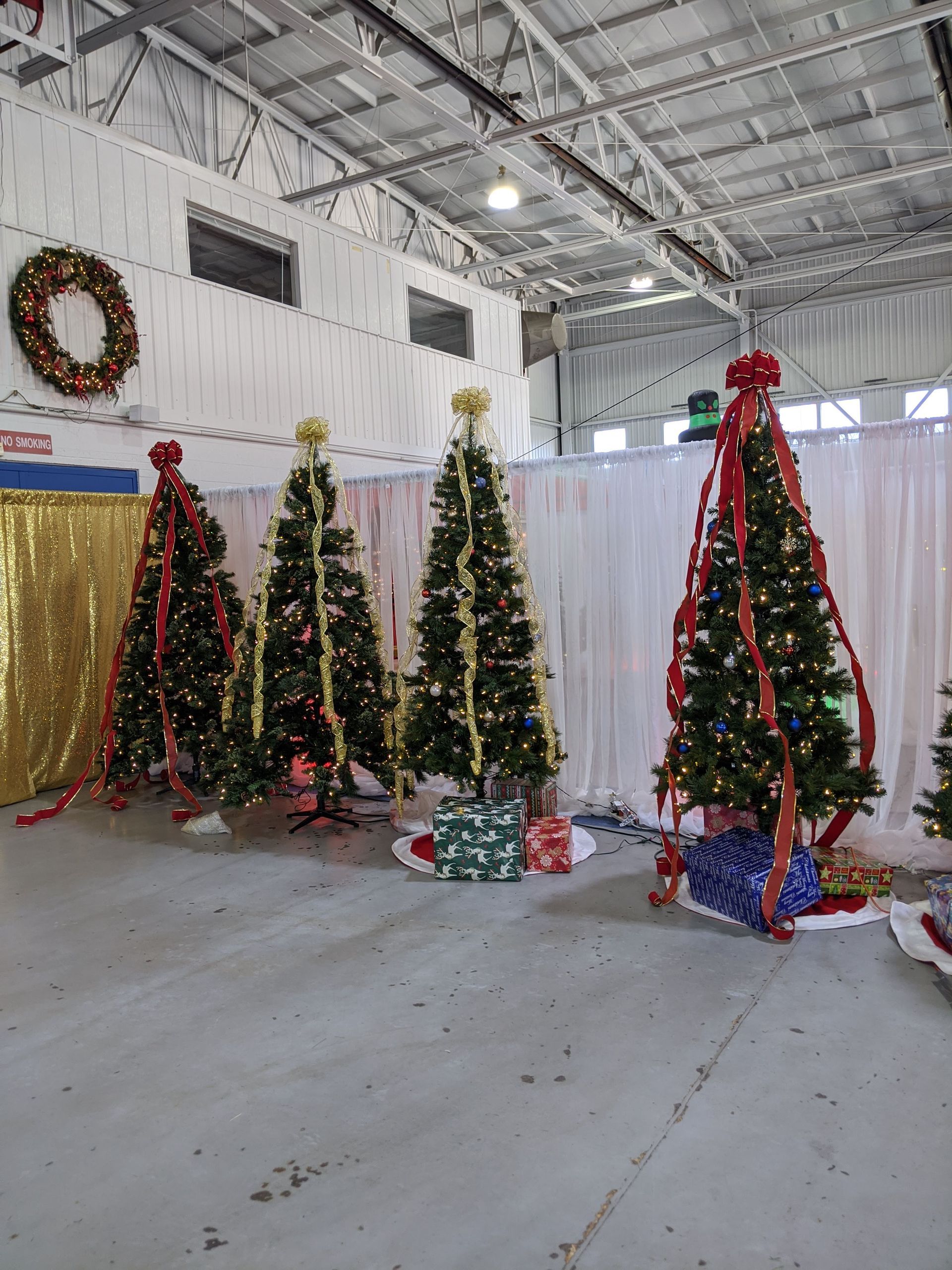 Decorated Christmas trees displayed in a large indoor hall with a concrete floor and holiday wreaths on the wall