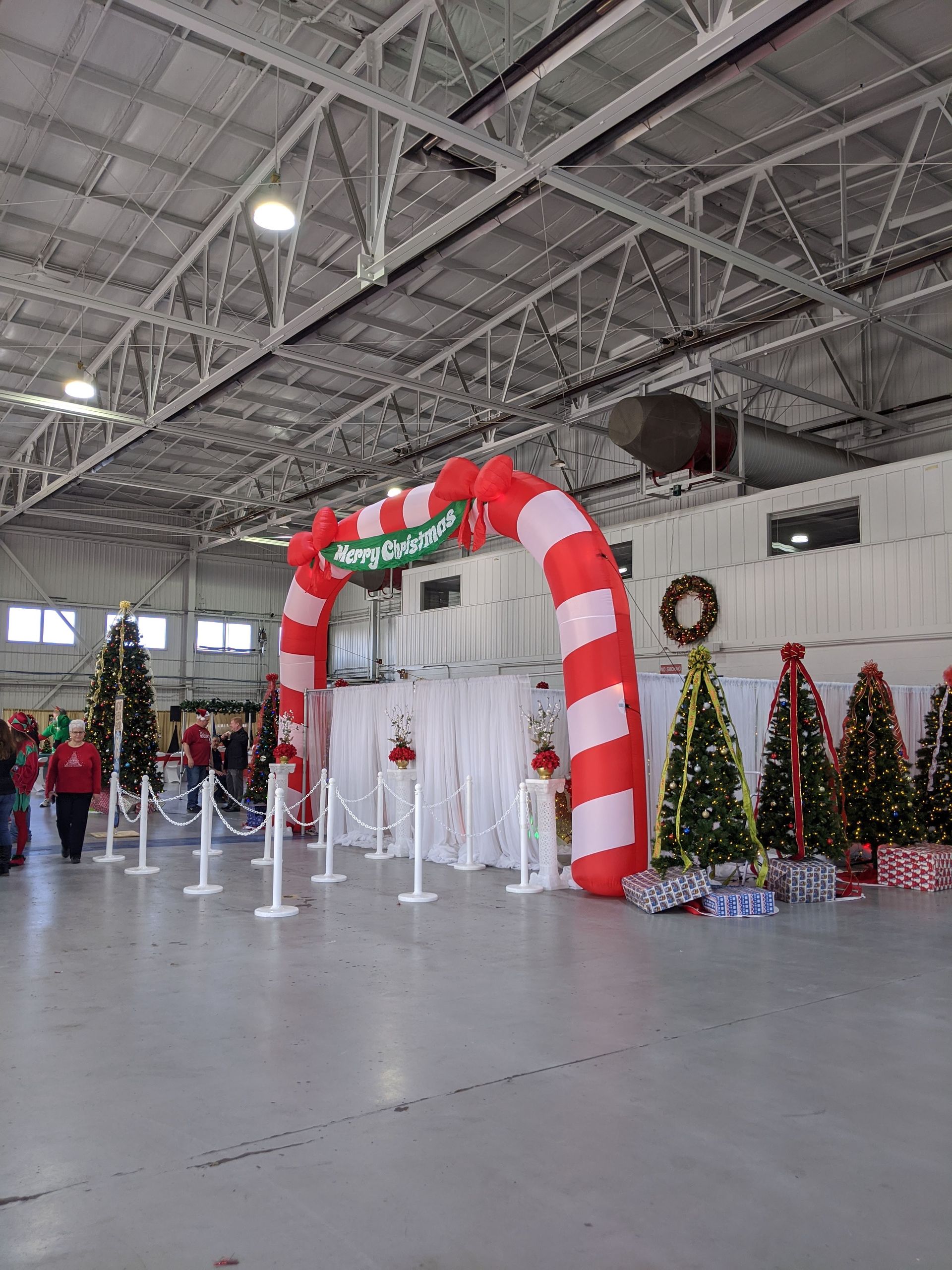 Indoor holiday display with red-and-white inflatable arch, Christmas trees, and Santa figures in a large hall