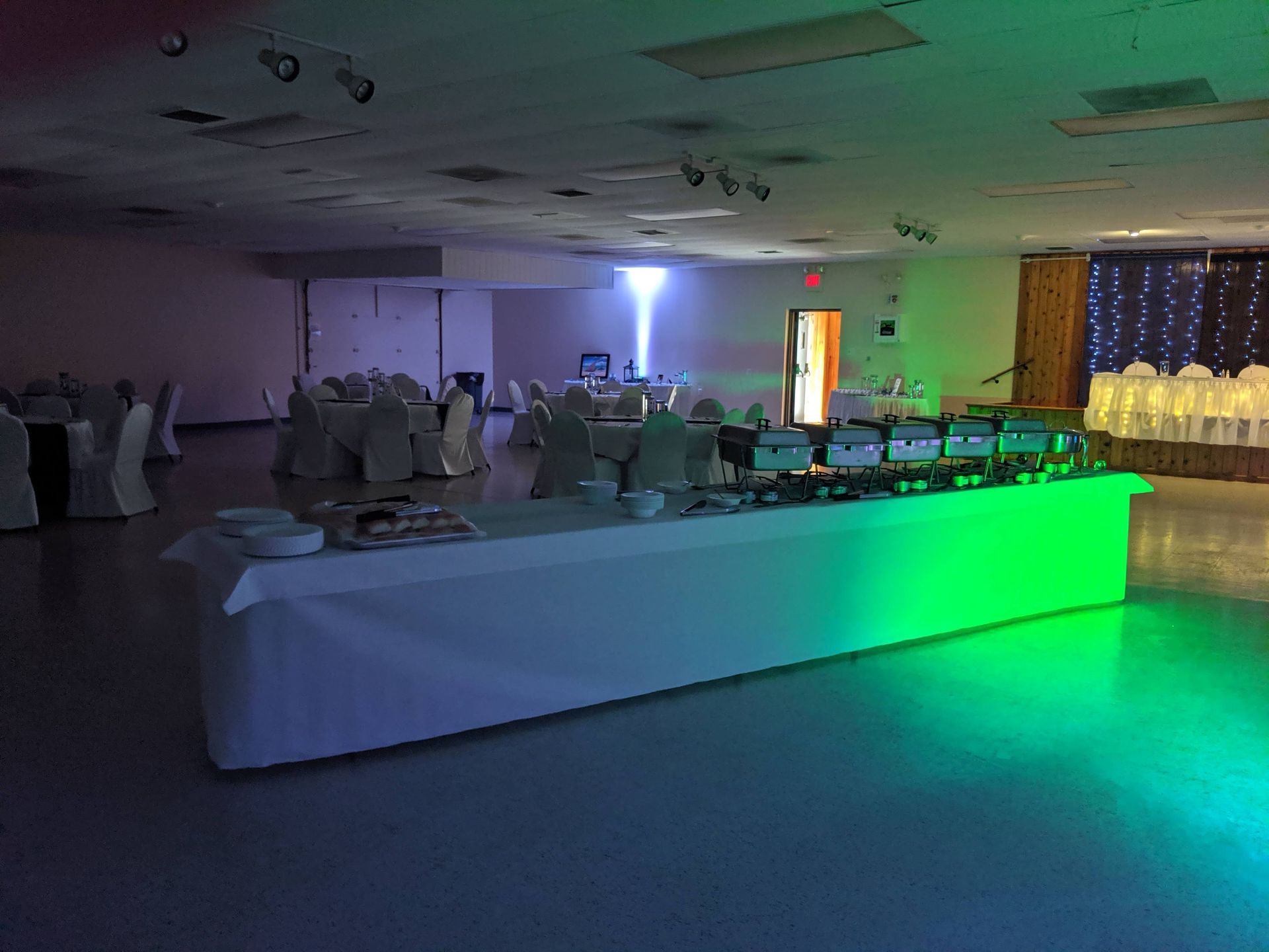 Dim banquet hall with round tables, white tablecloths, and green-blue uplighting near a dance floor.