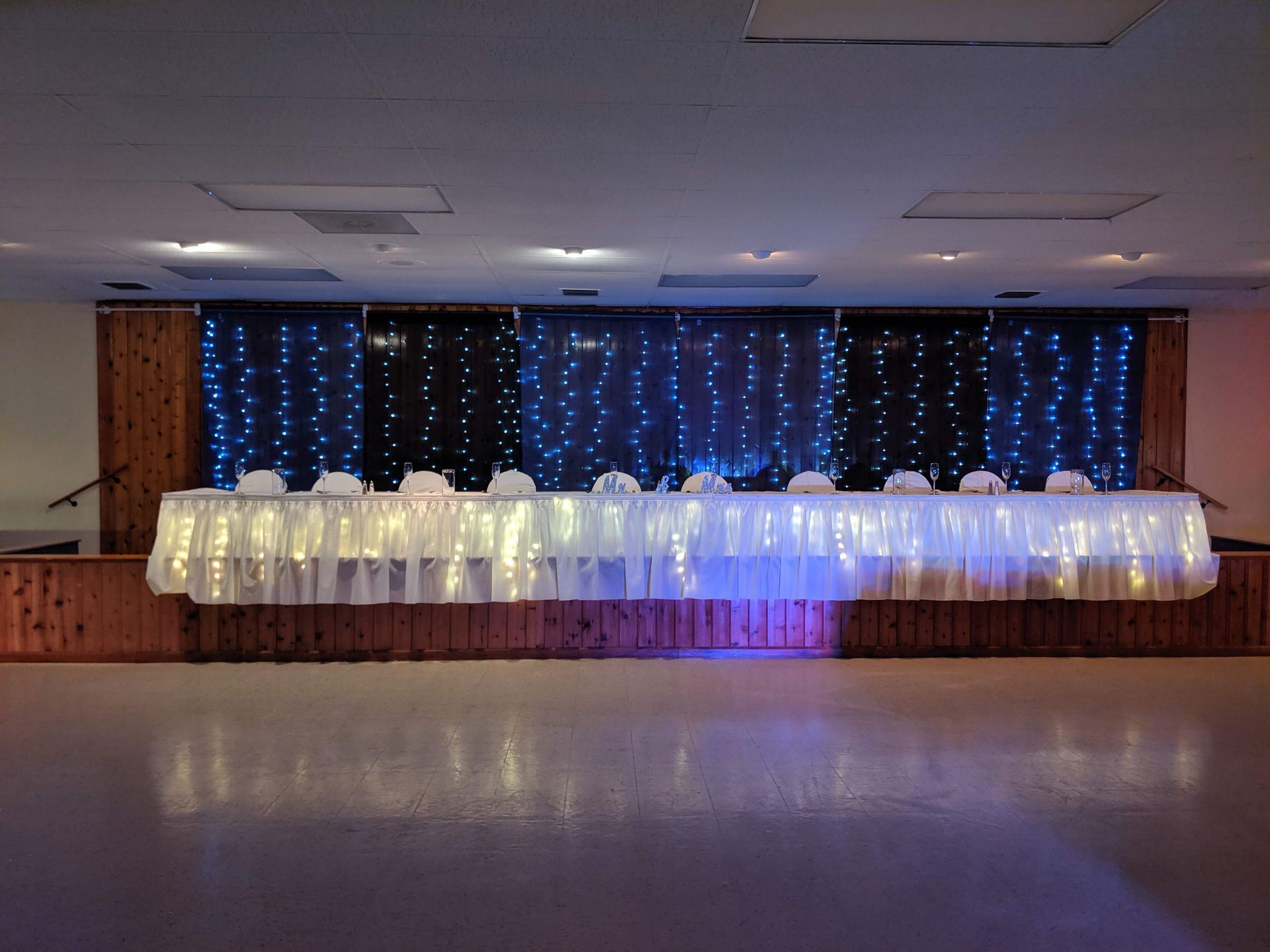 Long banquet table with white tablecloth and blue fairy lights on a stage in a hall