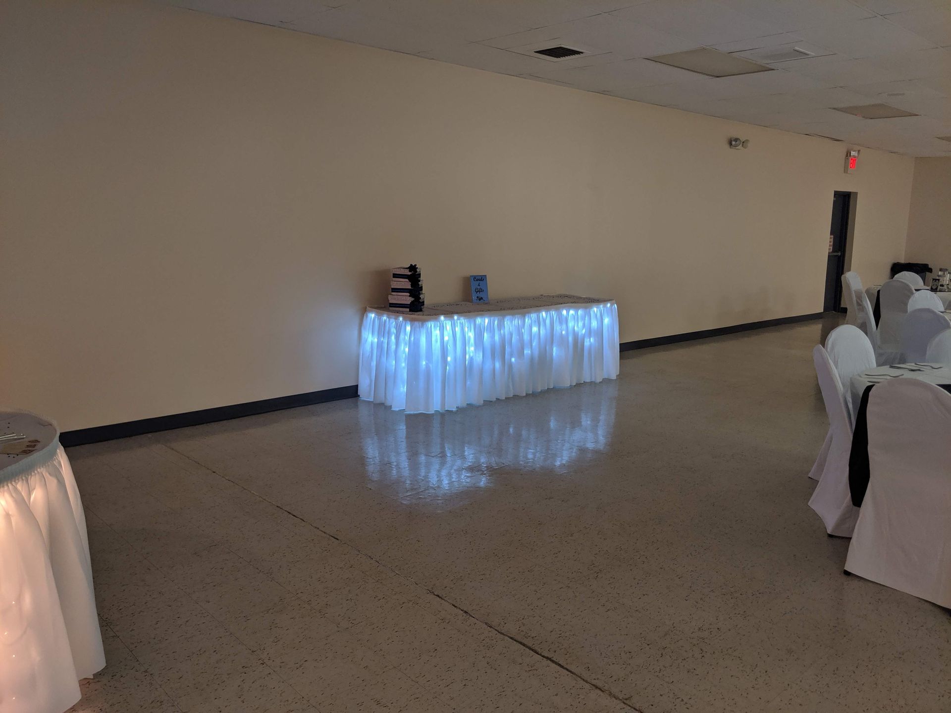 Empty banquet hall with illuminated white draped table at the back and round tables along the side стенов