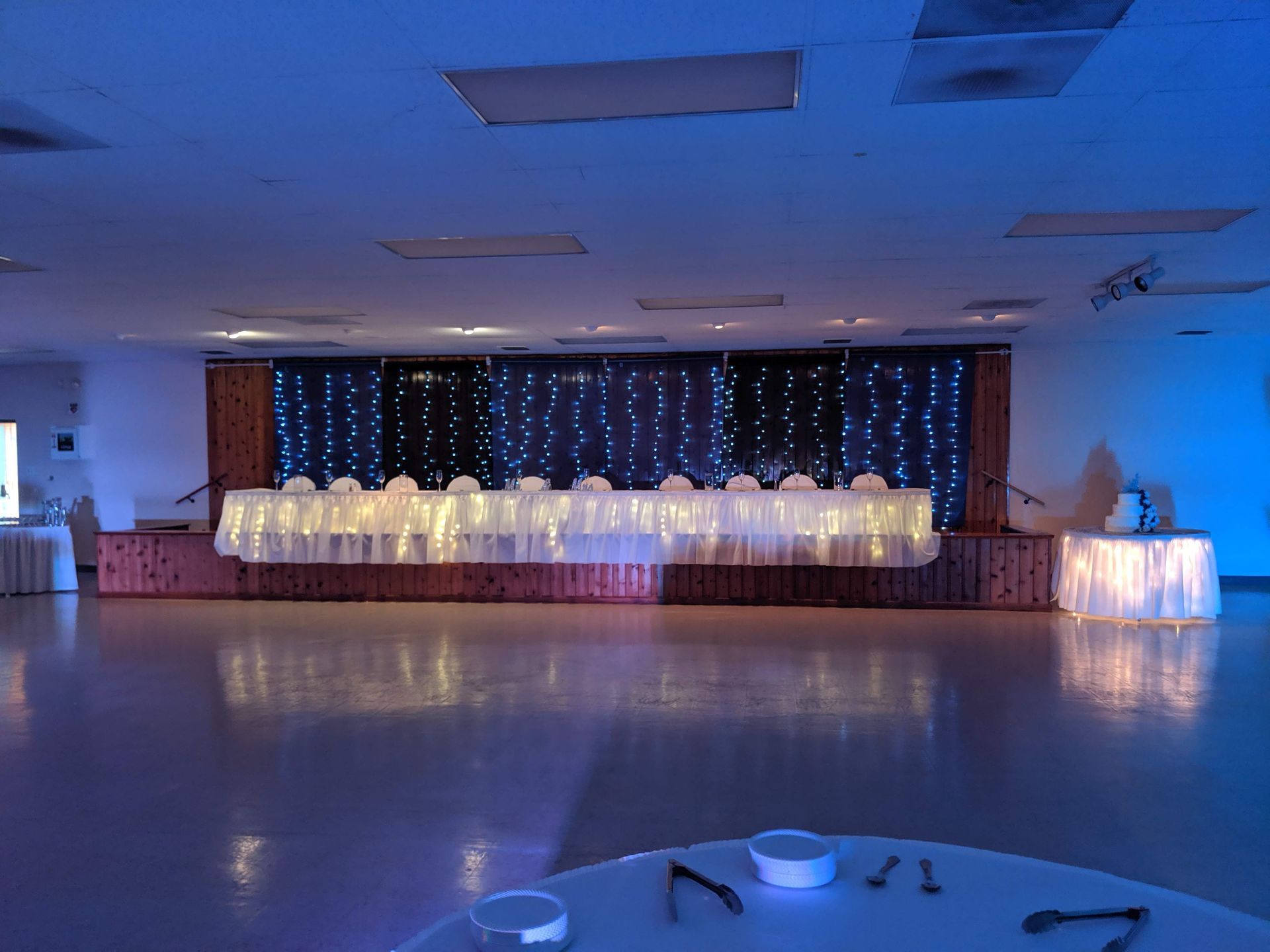 Blue-lit banquet hall with a long decorated head table and string lights along the back wall
