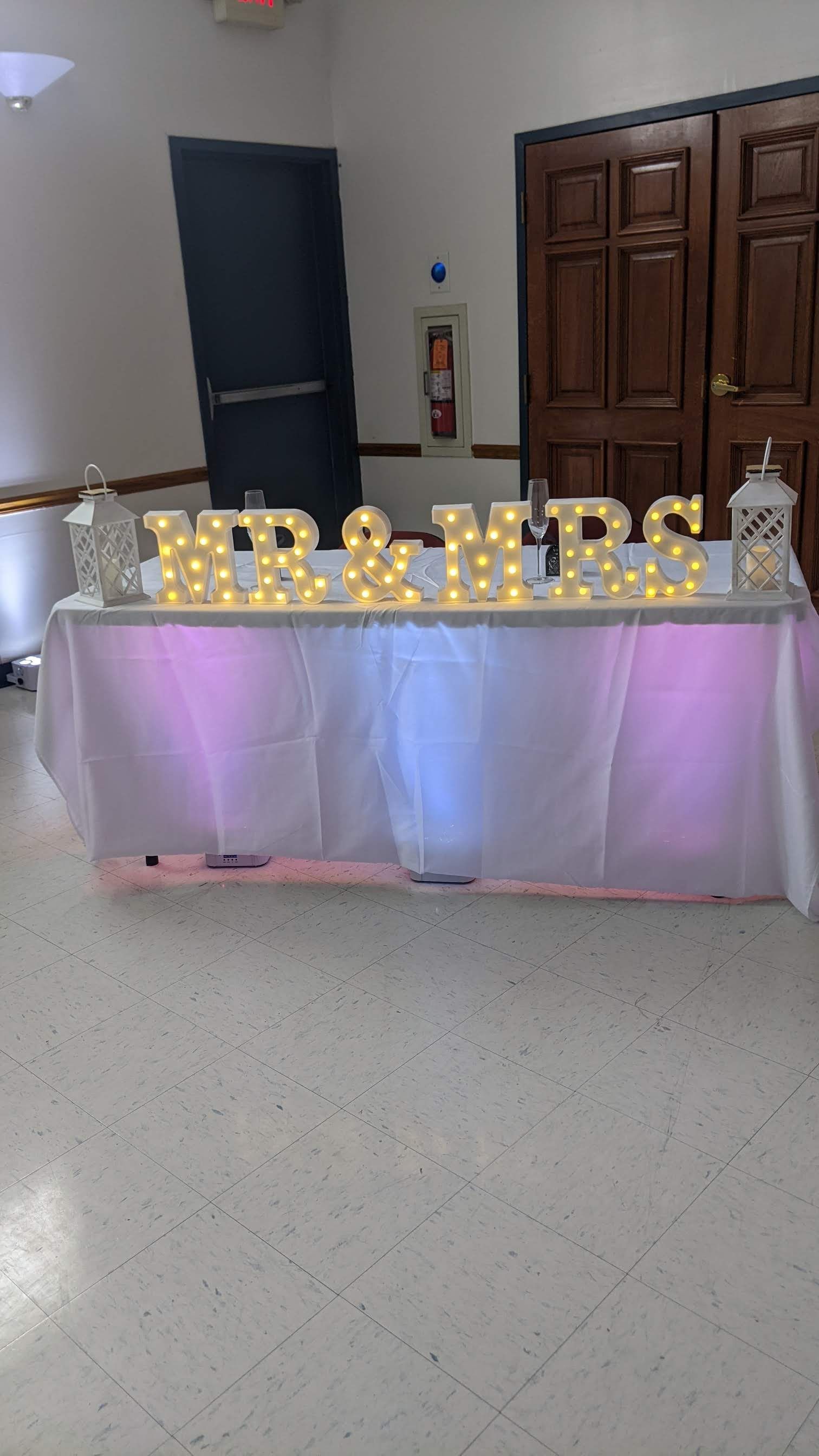 Decorated table with gold “MRS & MRS” letters and pink-purple lighting at an indoor event venue