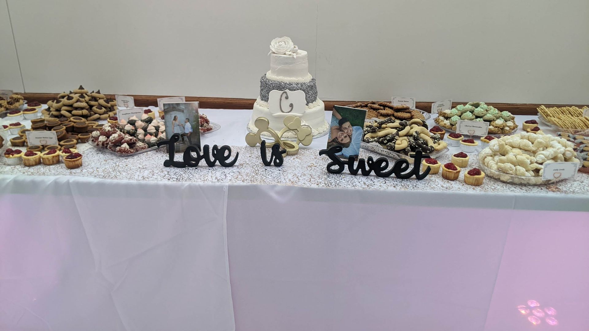 Dessert buffet with a white tiered cake and sweets, decorated with “love sweet” lettering on a white tablecloth.