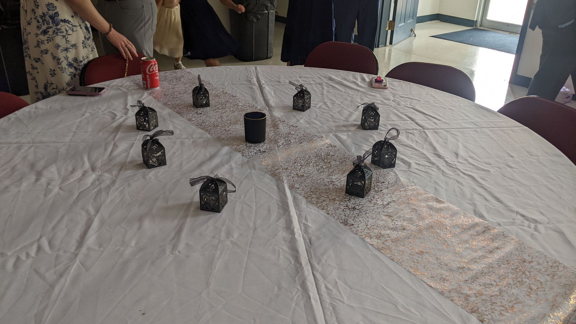 Small black objects arranged in a loose circle on a white tablecloth in a room with chairs and people nearby
