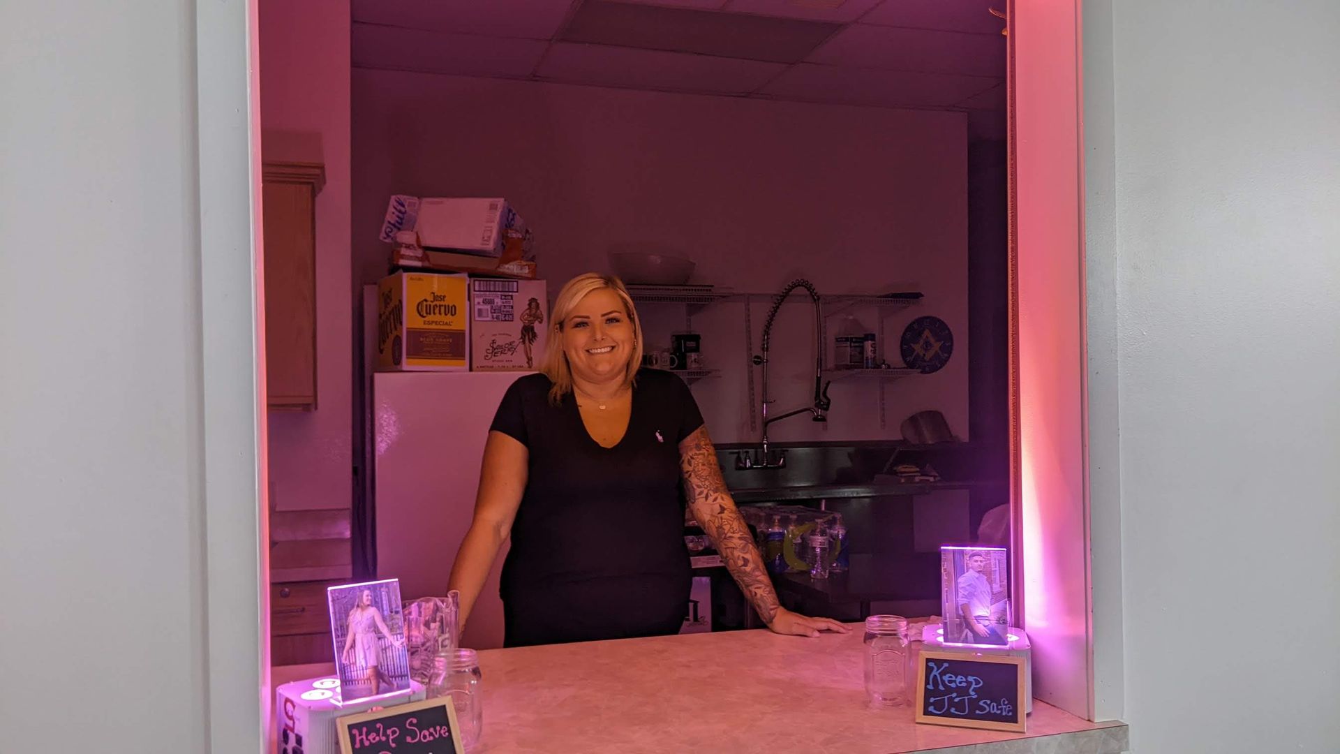 Woman standing behind a counter in a pink-lit room with glowing decorations and a kitchen in the background
