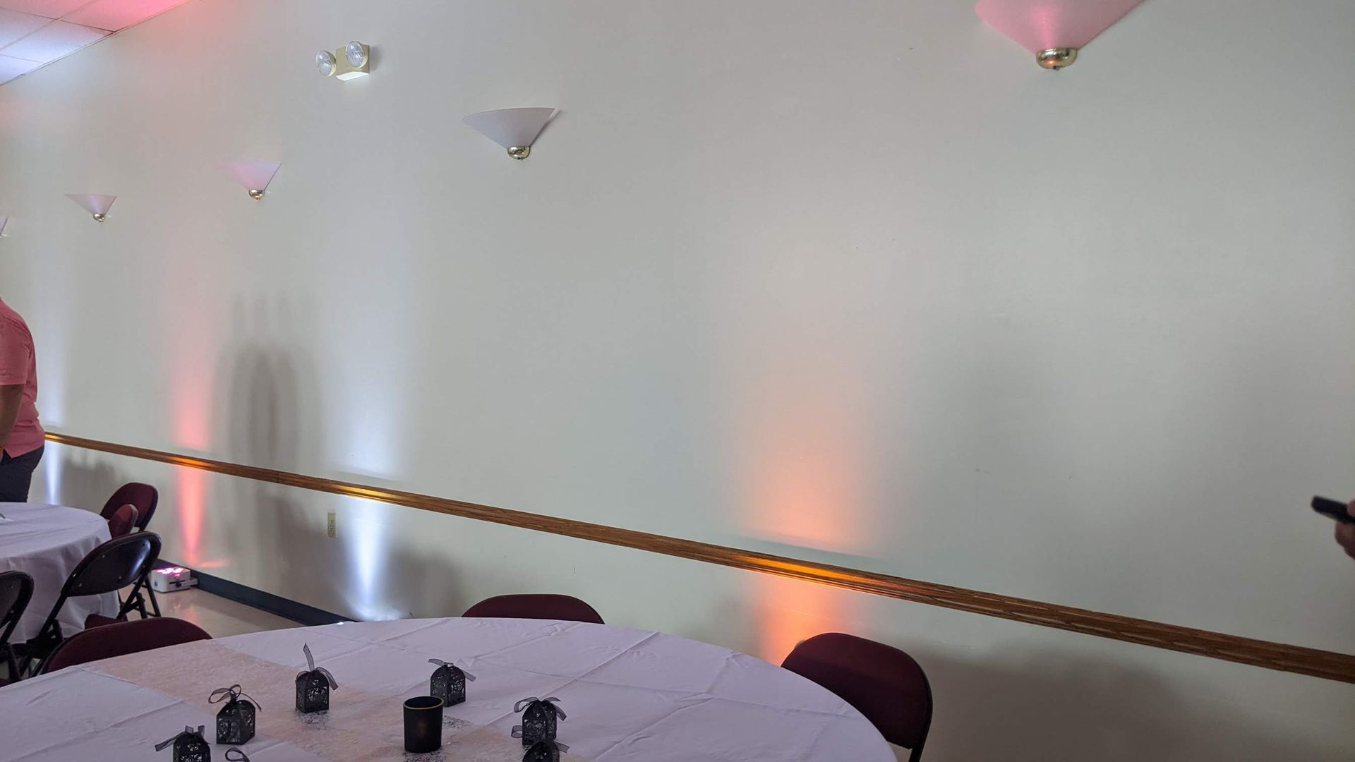 White event room with round tables, maroon chairs, wall sconces, and a person at left near the aisle