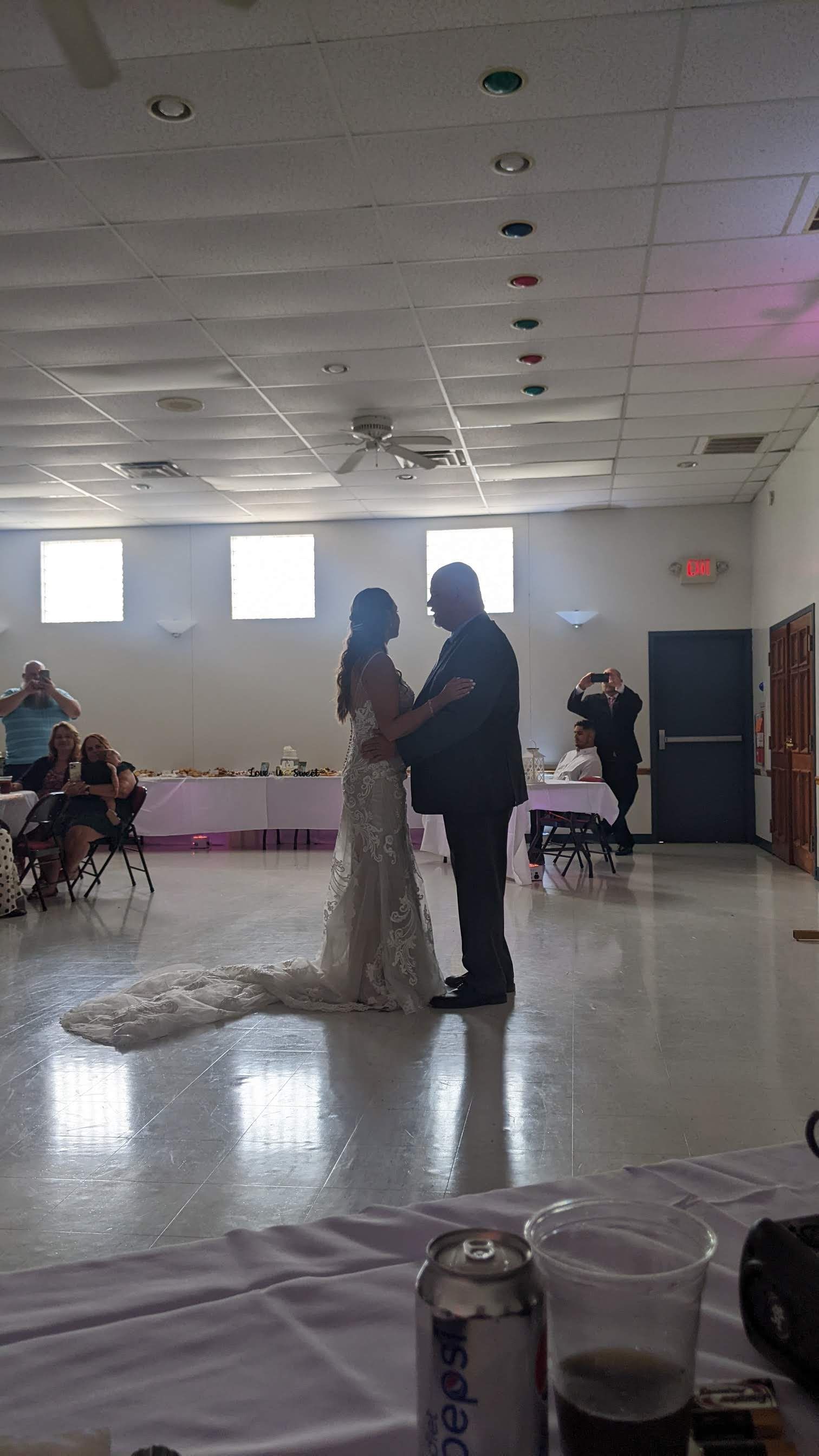 Couple slow dancing at a reception hall, with guests seated around the room and soft ceiling lights above