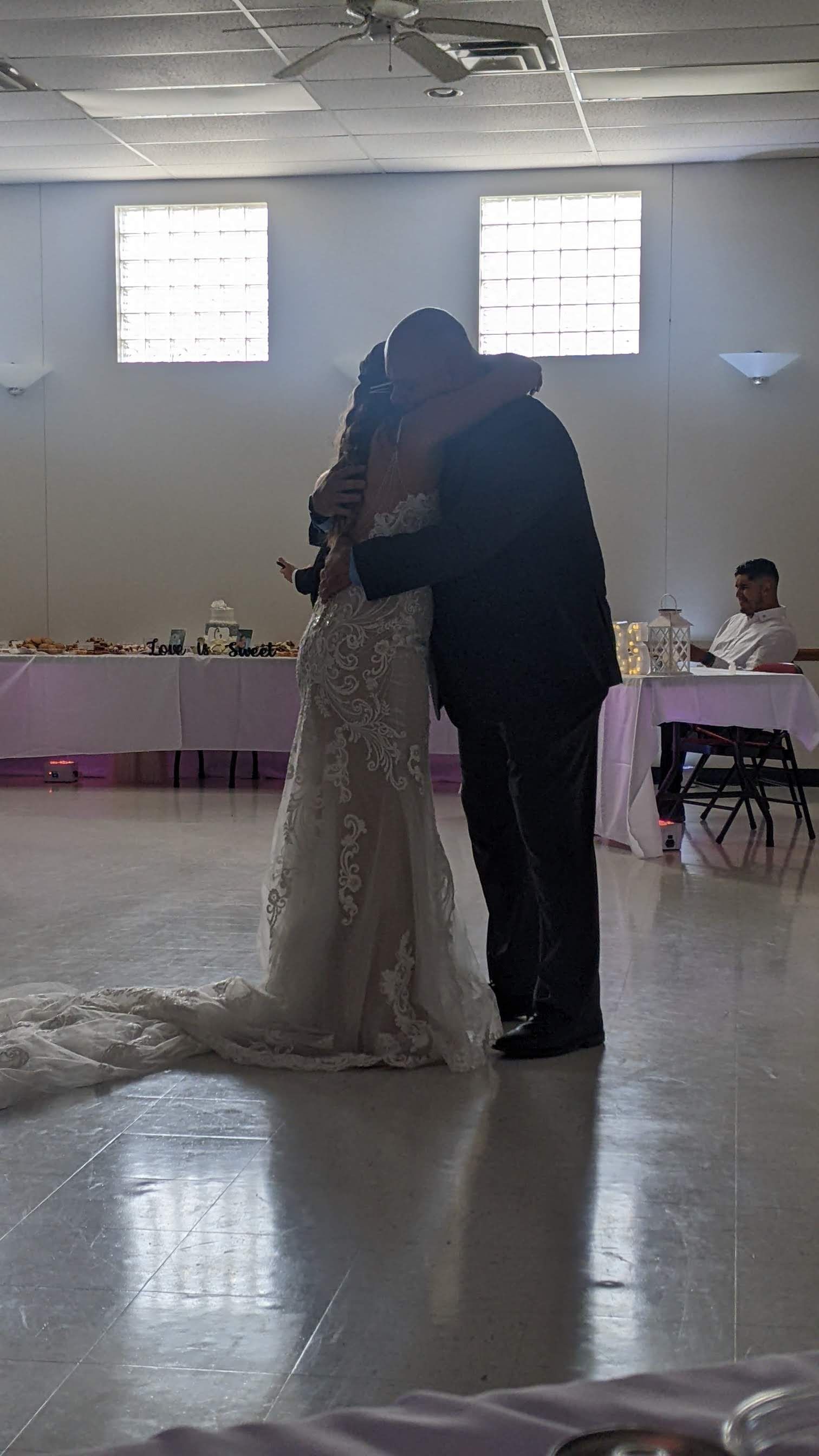 Couple slow dancing at a wedding reception in a bright hall with guests seated nearby.