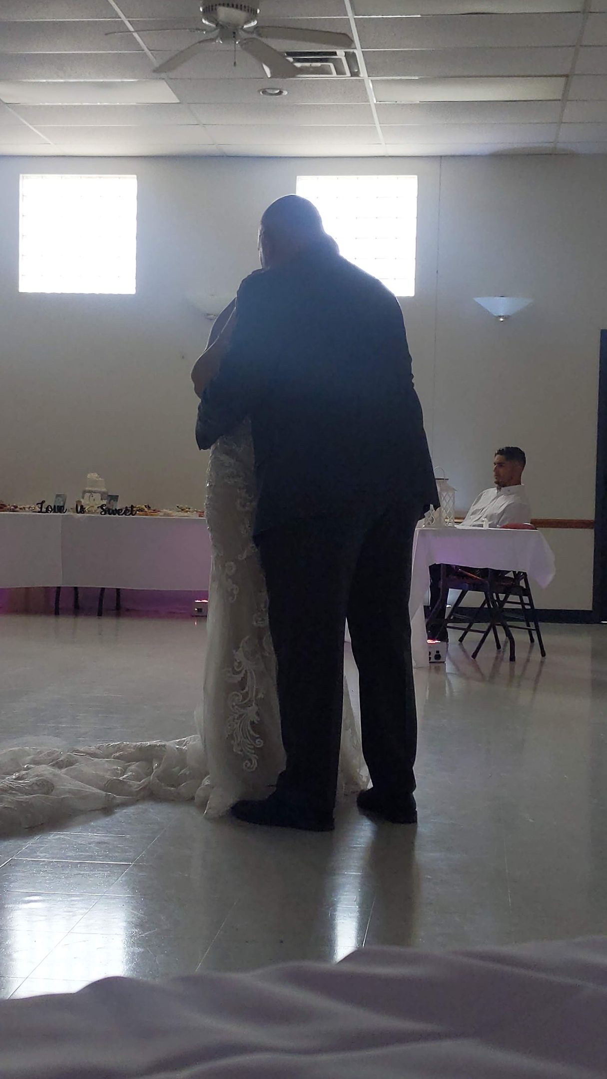 Two people standing and embracing in a bright indoor hall with white walls and a tiled floor.
