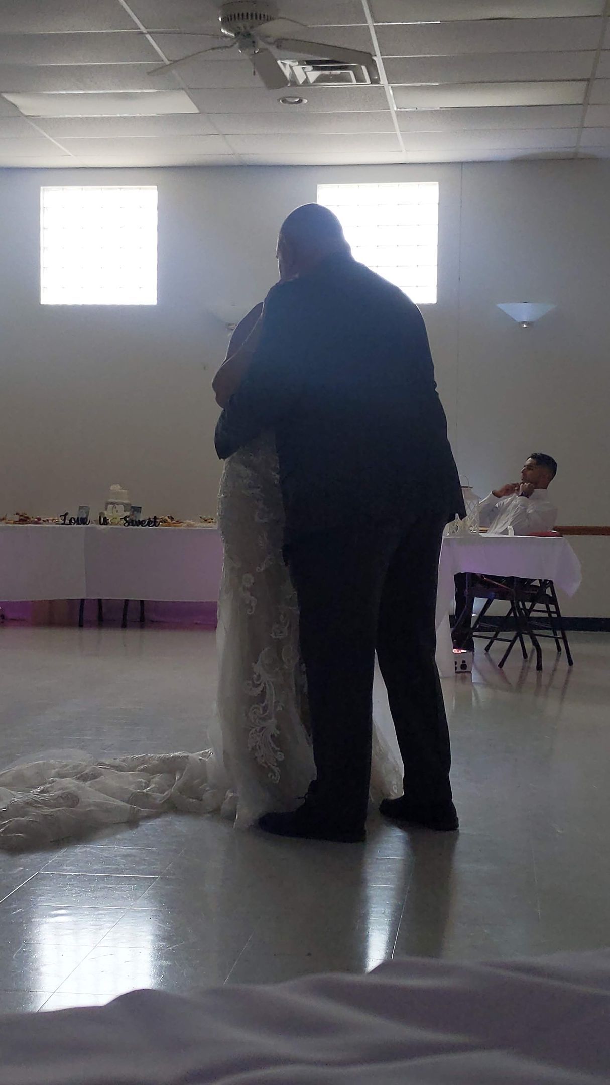 Two people standing and embracing in a dim hall with a reflective floor and bright windows.