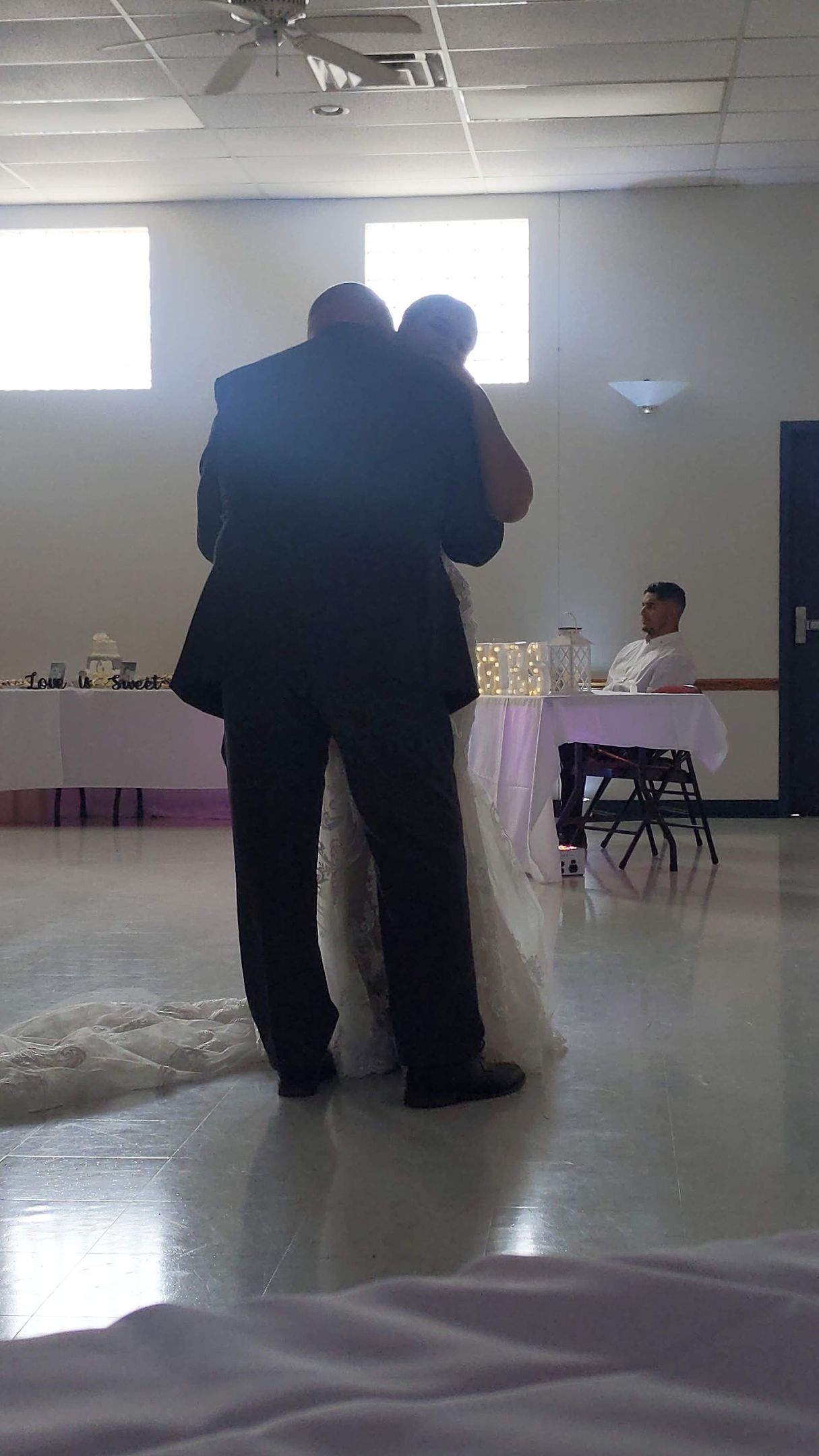 Two people hugging in a bright room with tables and chairs in the background.