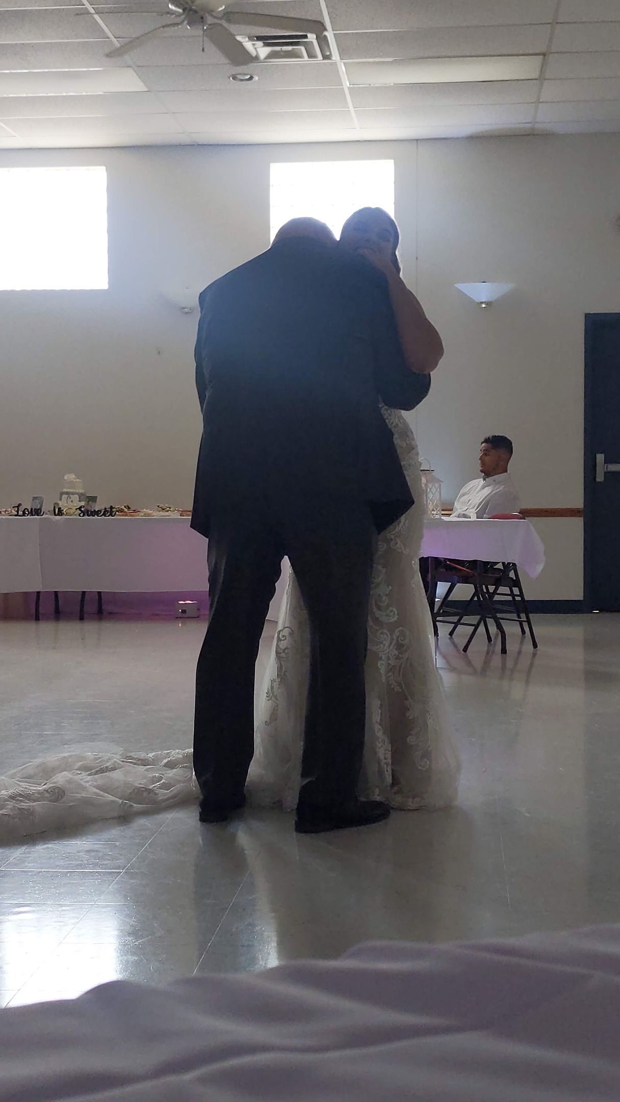 Two people embracing in a bright hall with tables and seated guests in the background.