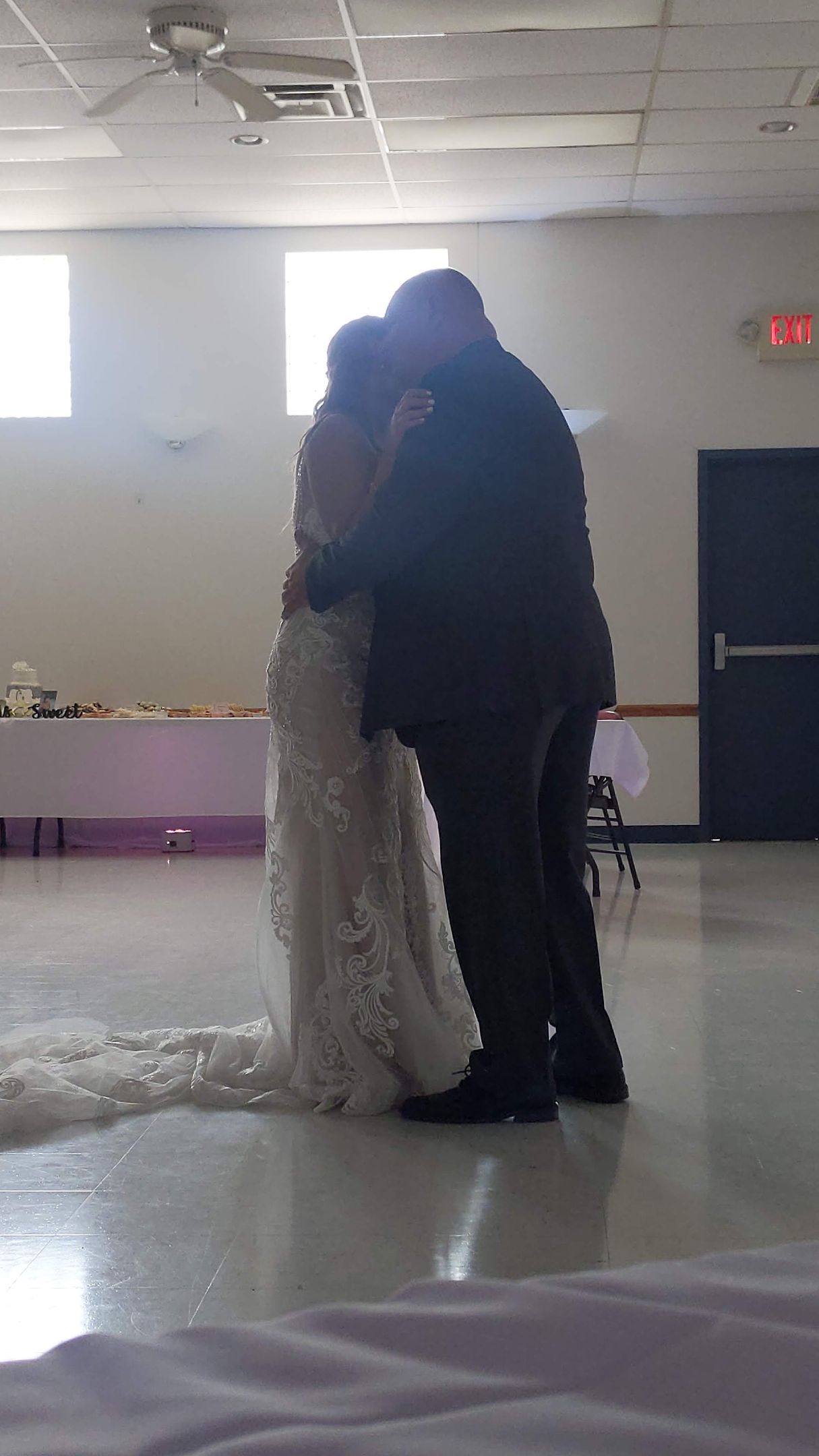 Two people in formal attire embrace during a dance in a bright indoor hall.
