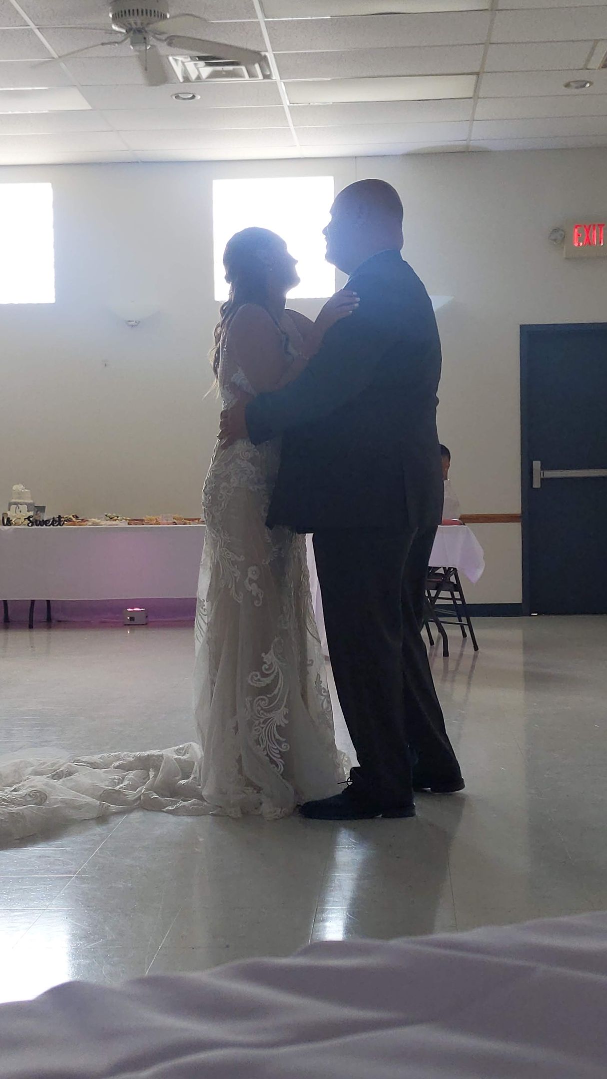 Two people dancing close together in a dim indoor hall with white walls and a tiled floor.