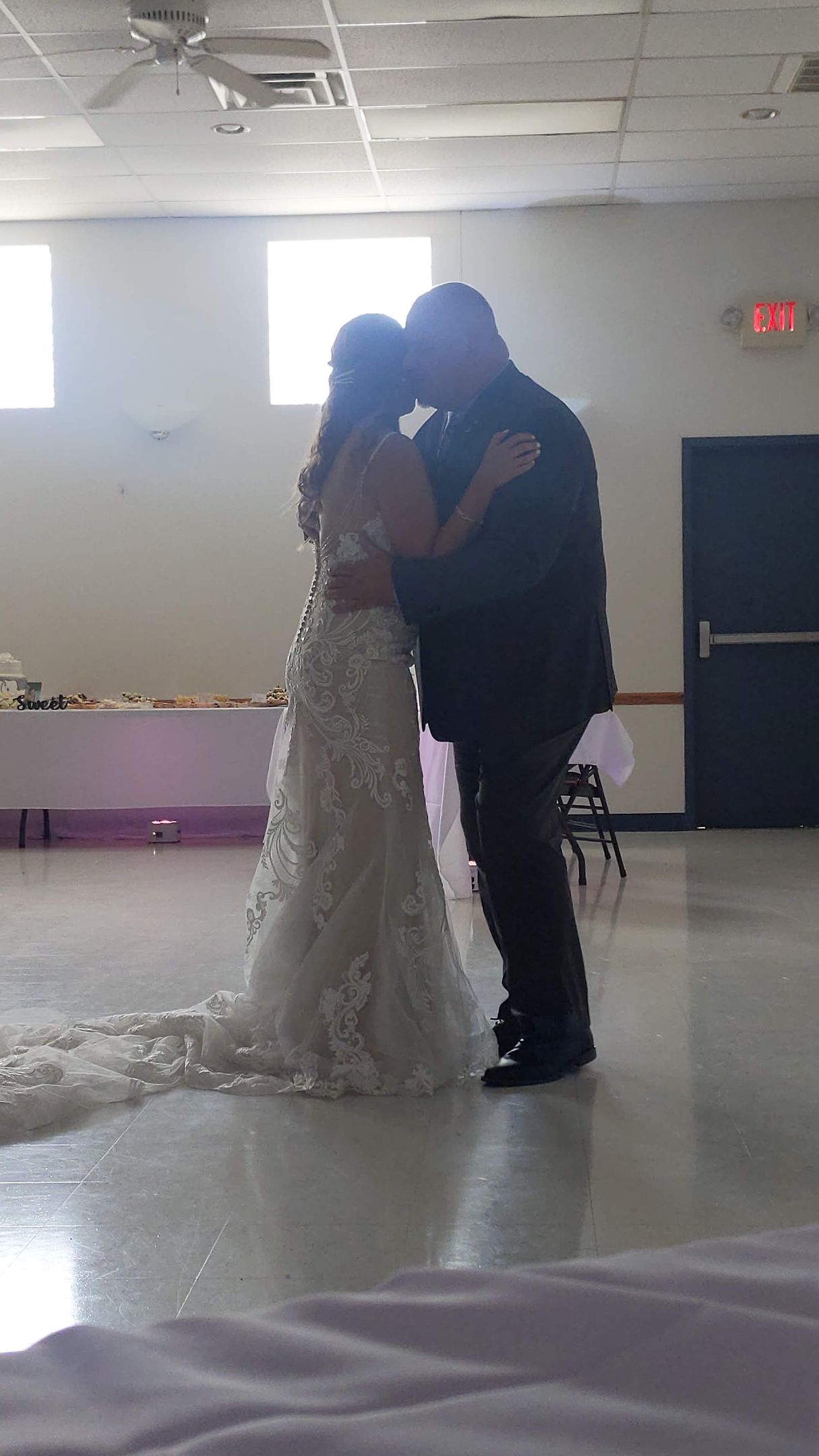 Two people in formal attire sharing a dance in a bright room with white walls and purple table drapes.