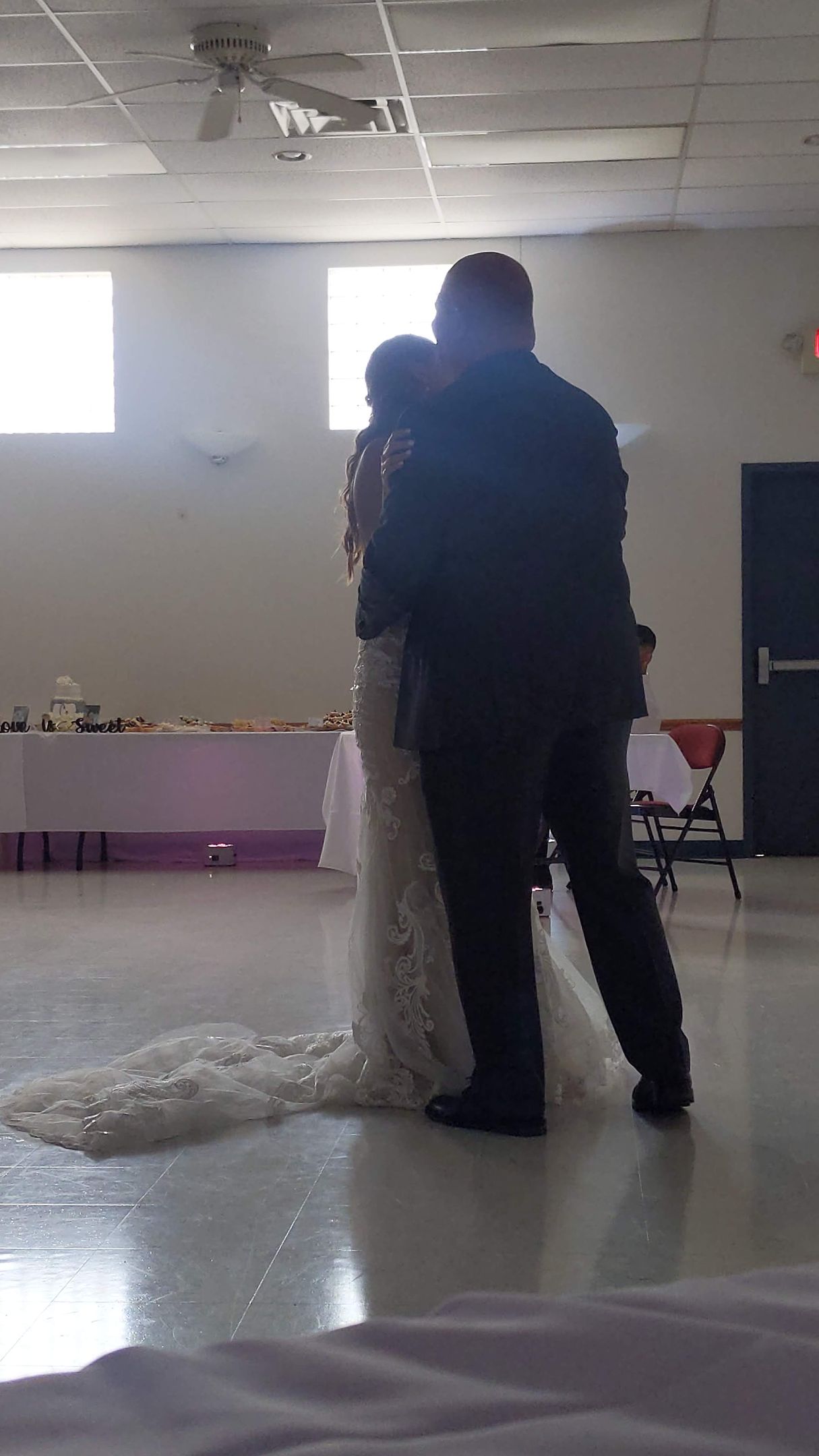 Two people embrace in a bright hall, with a white draped stand beside them and flowers in the background.