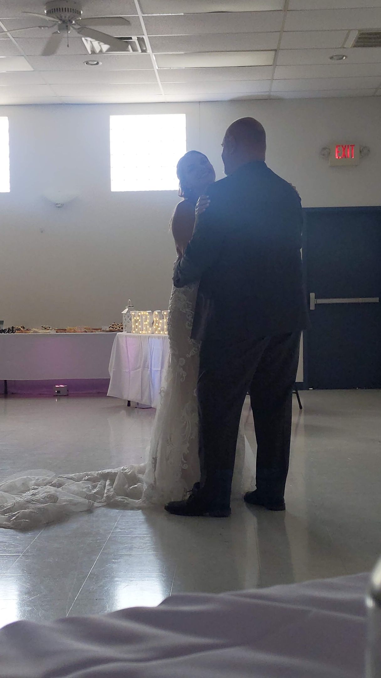 A couple slow dancing at a reception, silhouetted near a bright window in a hall.