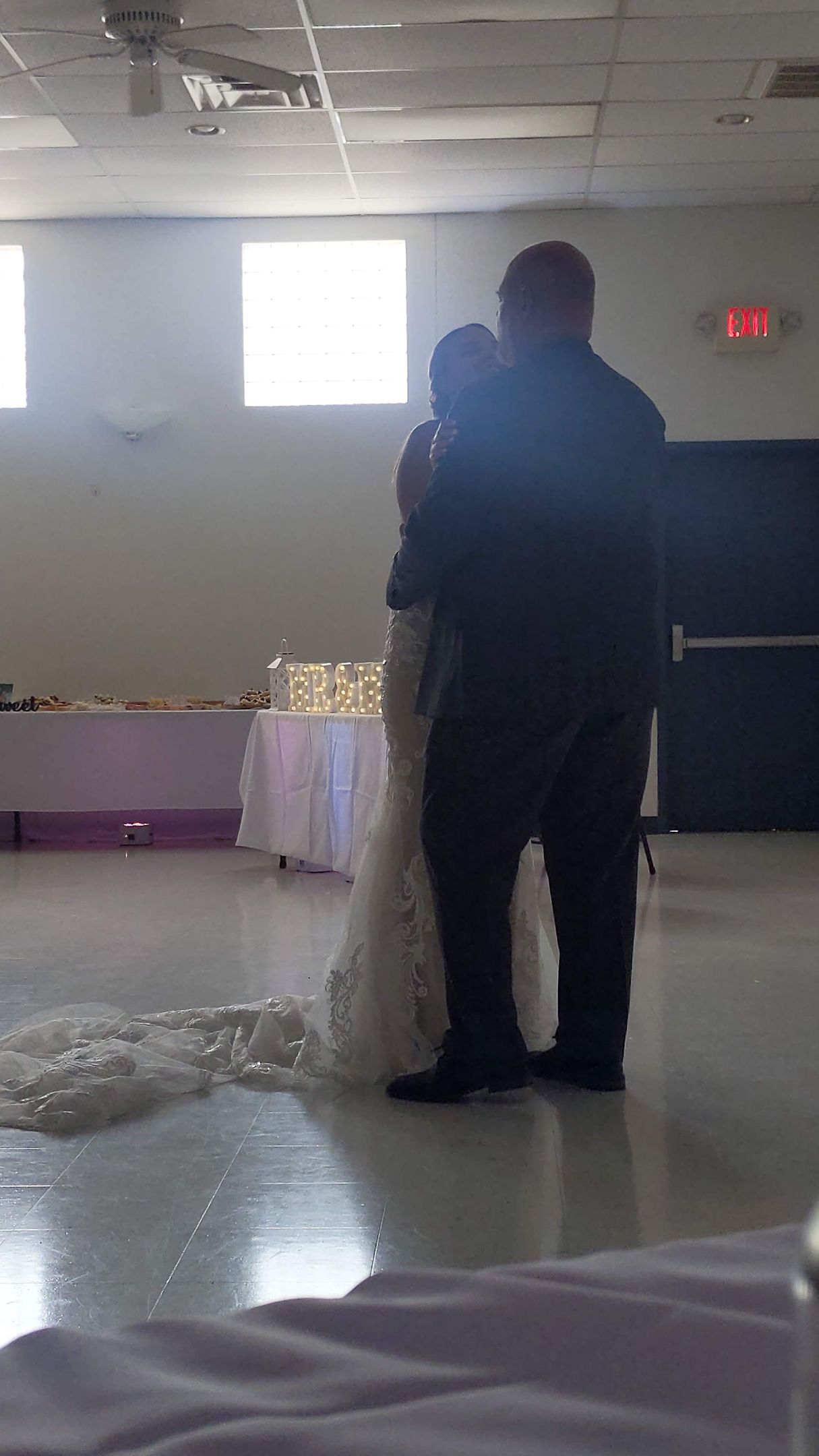 Person in dark clothing standing in a hall with a child in a white dress during a formal indoor event.