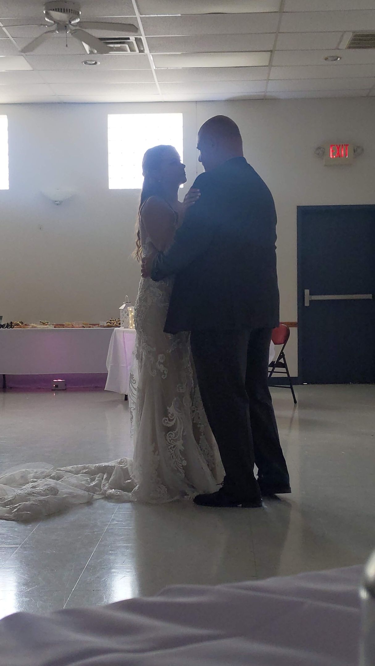 Two people embracing in a dim hall, one in a white lace gown and one in a dark suit.