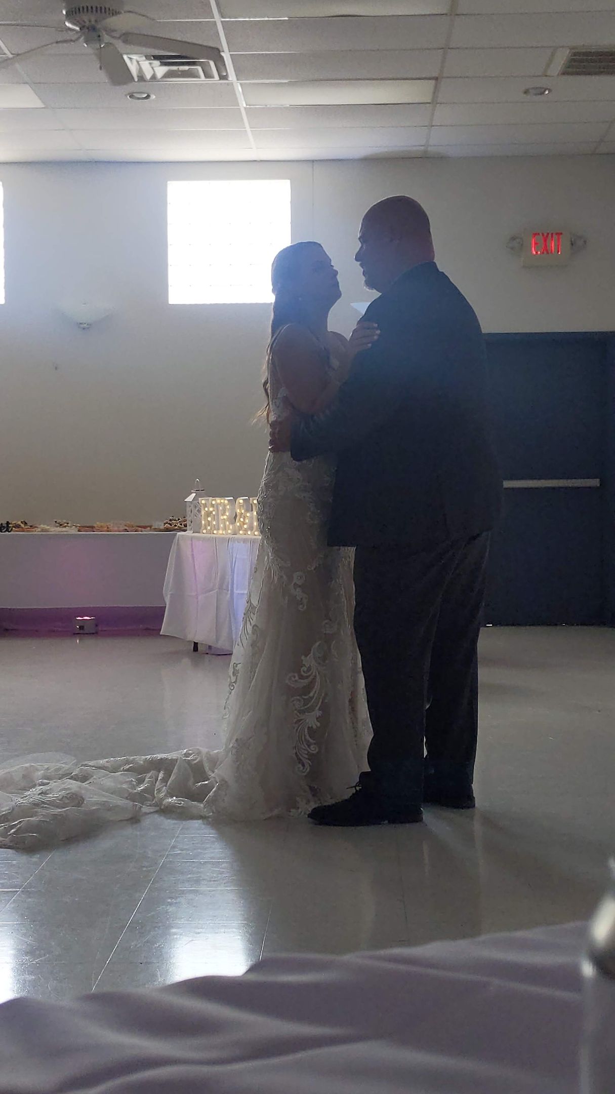 Two people slow-dancing in a dim hall, with a white table and bright window in the background.