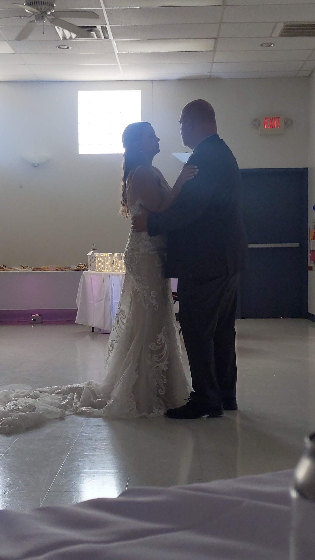 Bride and groom stand facing each other at a wedding reception, silhouetted by bright backlight.