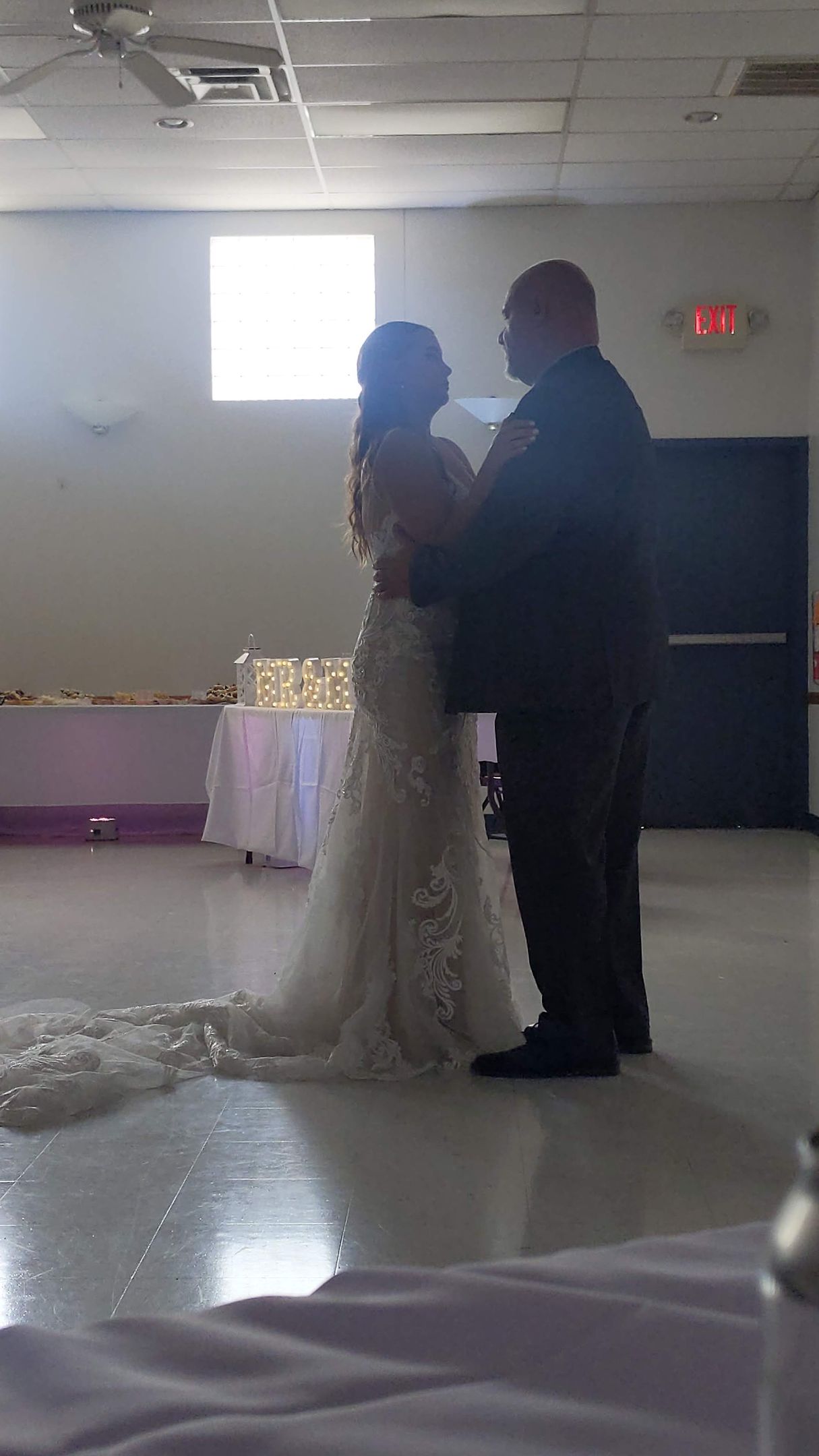 Bride and groom dancing in a banquet hall with a bright window behind them