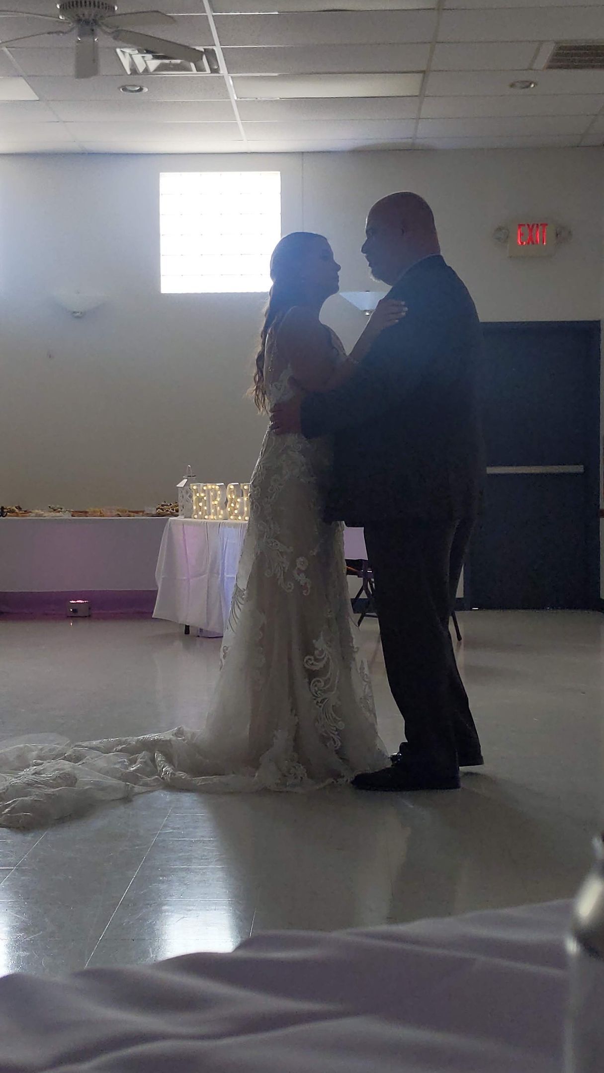 Two people dancing at a wedding reception in a decorated hall, with a bright window behind them.