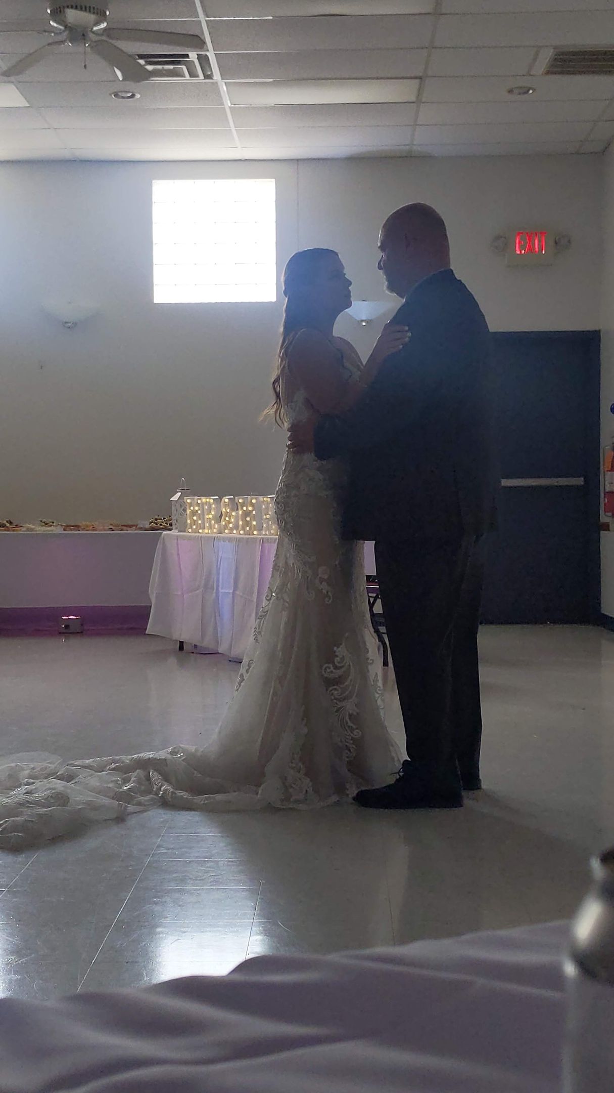 Bride and groom slow dancing at a dimly lit reception with a bright window behind them