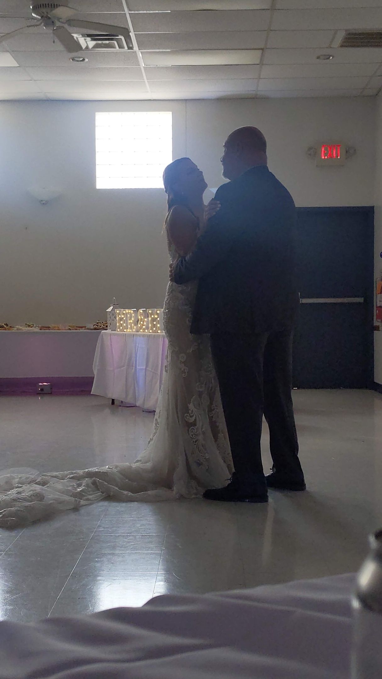 Two people stand together in a dim hall, one in a white dress, near a decorated table.