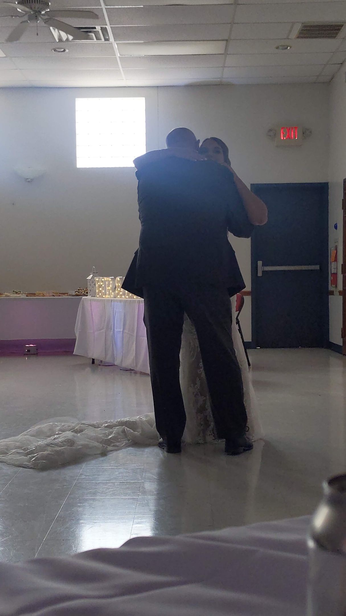Two people embrace on a polished floor in a hall with tables, a bright window, and a door in the background.