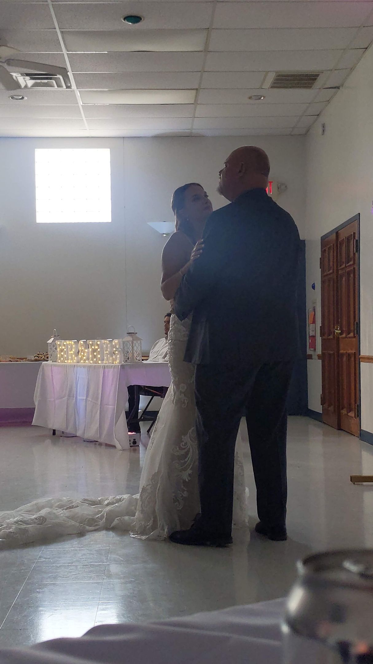 Two people stand in a hall beside a decorated table and white floral display beneath a bright window.