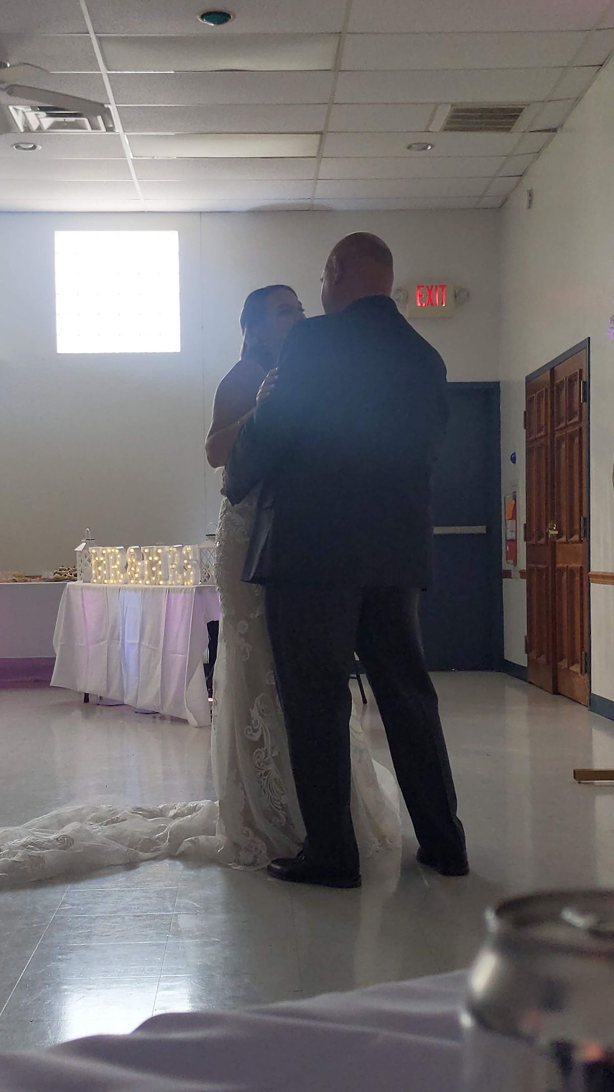 Two people standing together in a bright hall near a decorated table and exit sign.