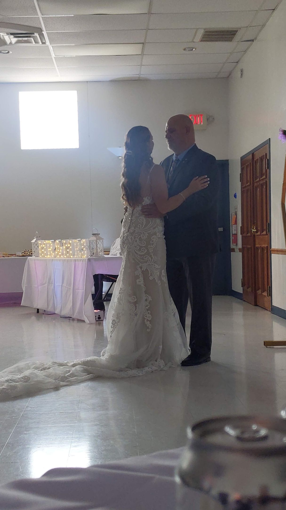 Bride and groom slow dance in a bright reception hall, bride in white gown and groom in dark suit.