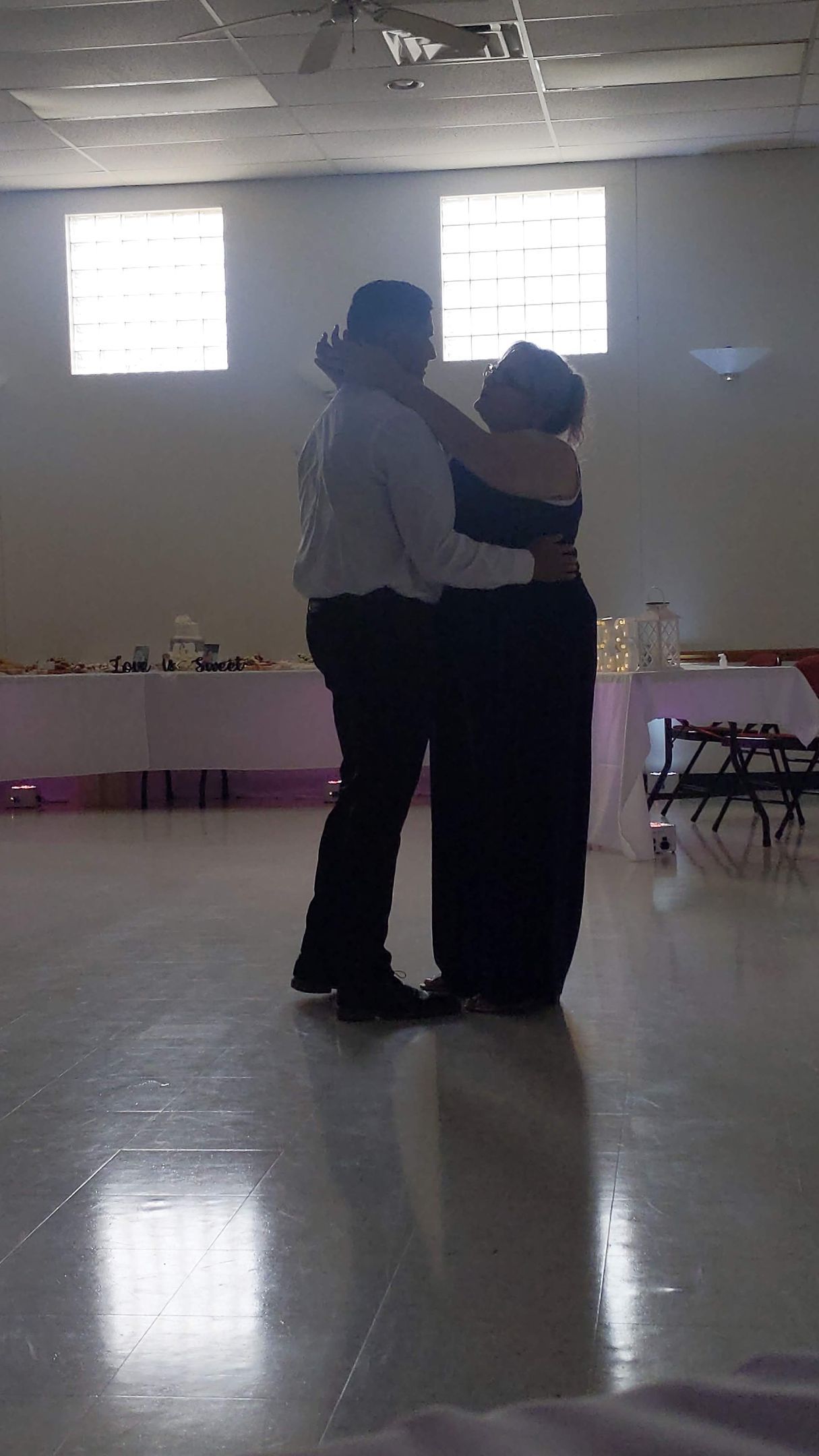 Two people dancing close together in a bright hall with high windows and a polished floor