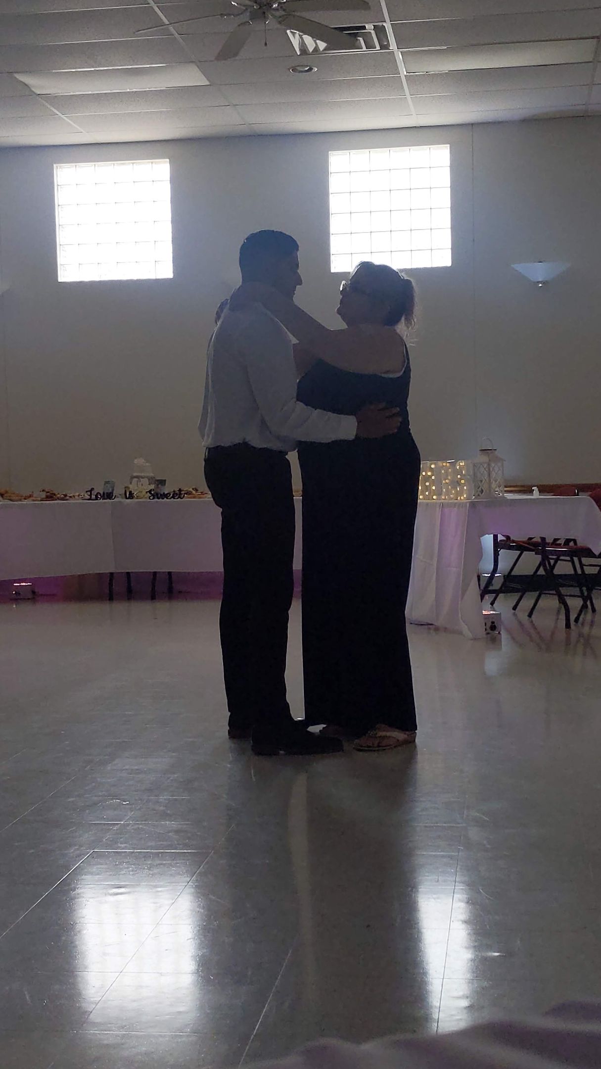 Two people embracing in a bright hall with windows and a reflective floor.