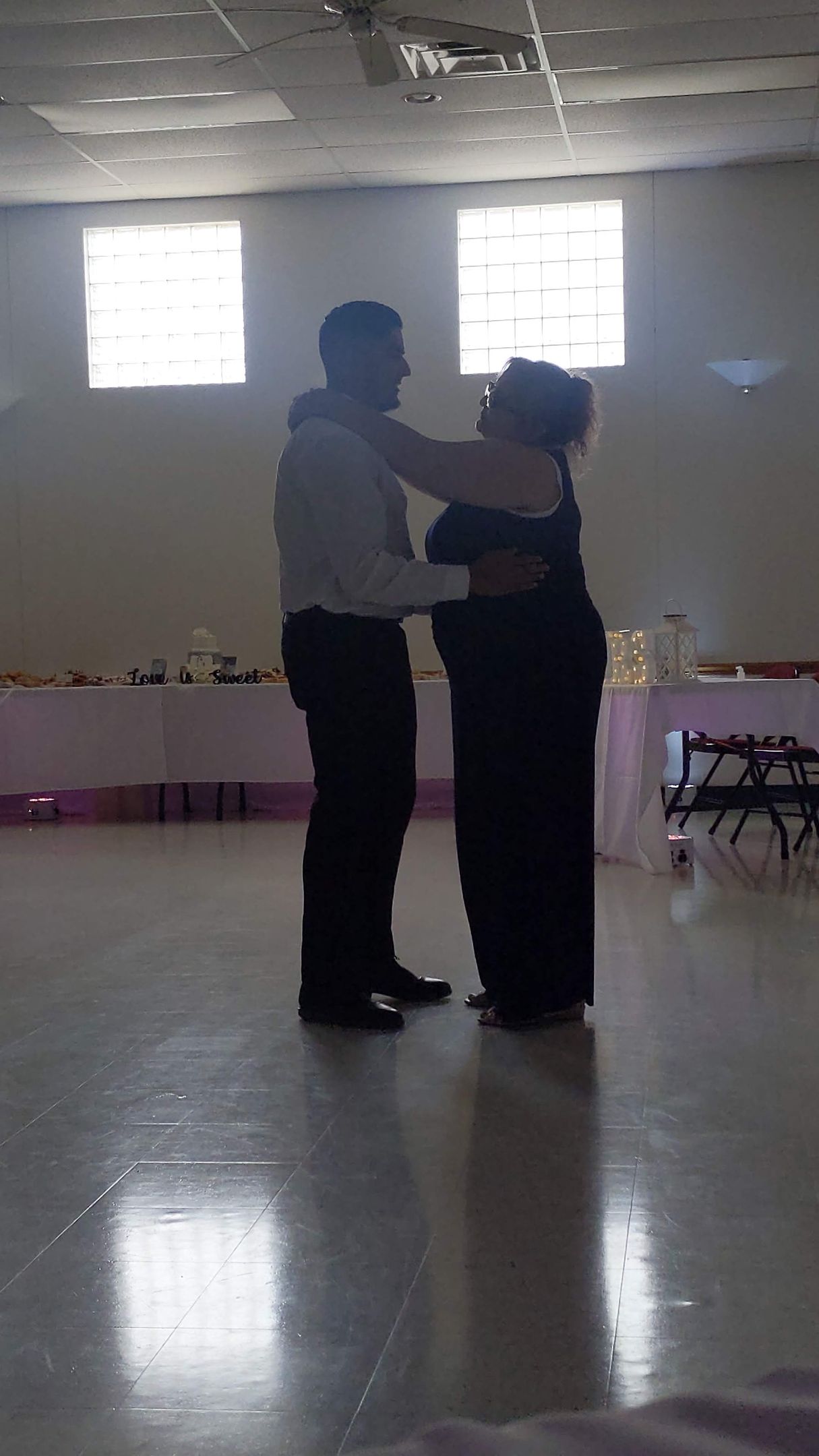 Two people dancing in a bright hall with high windows and a reflective floor.