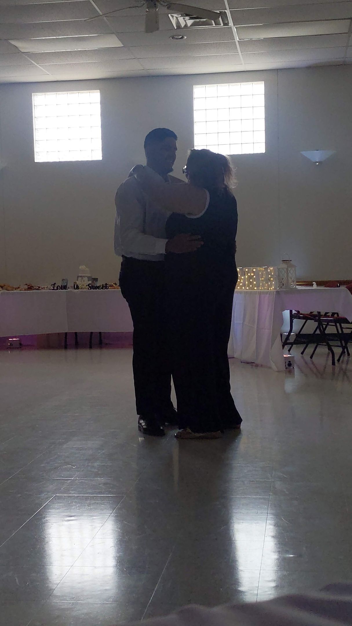 Two people dancing closely in a bright hall with reflective floor and tables along the wall.