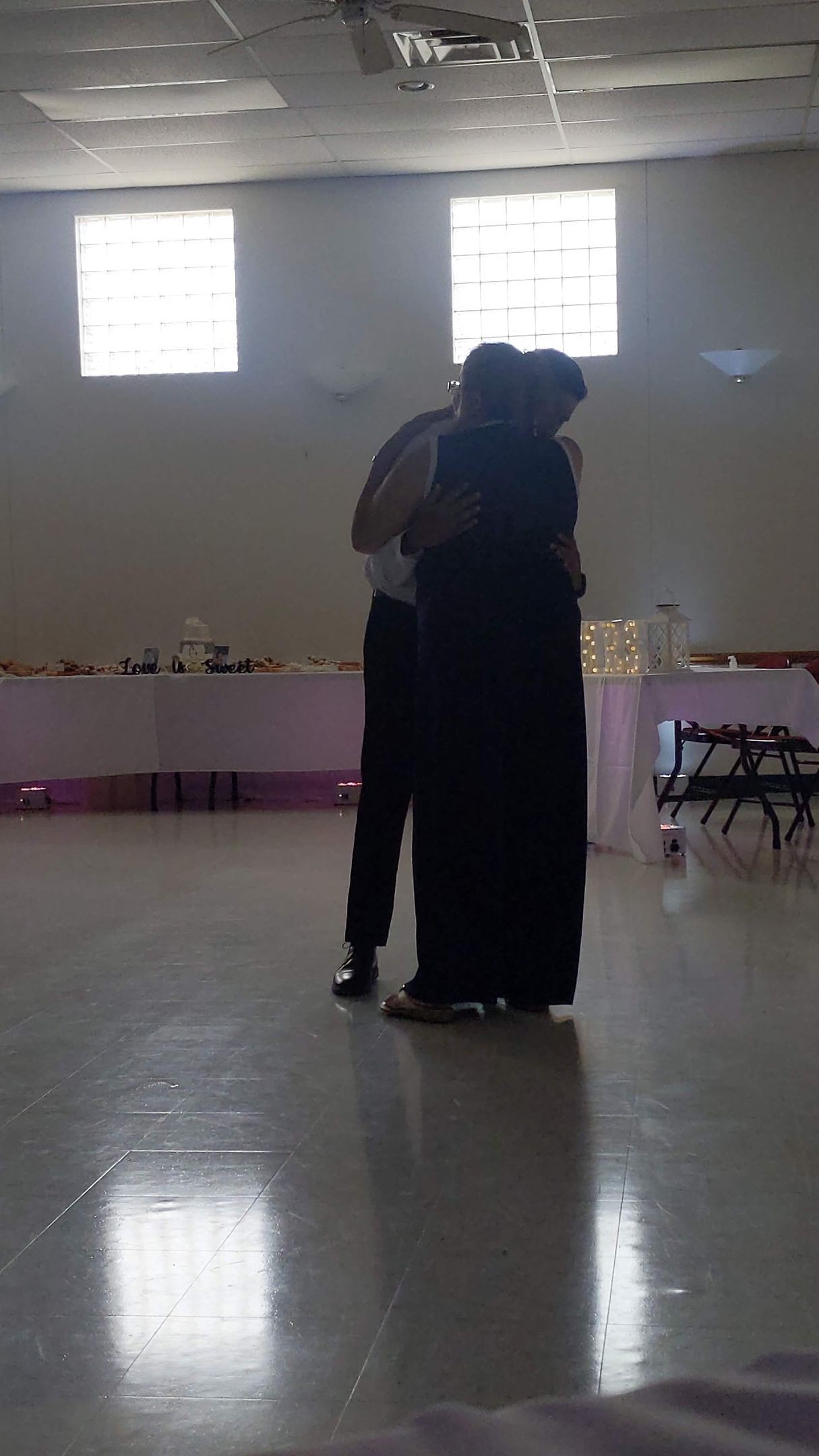 Two people embracing in a bright indoor hall with a reflective floor and windows behind them