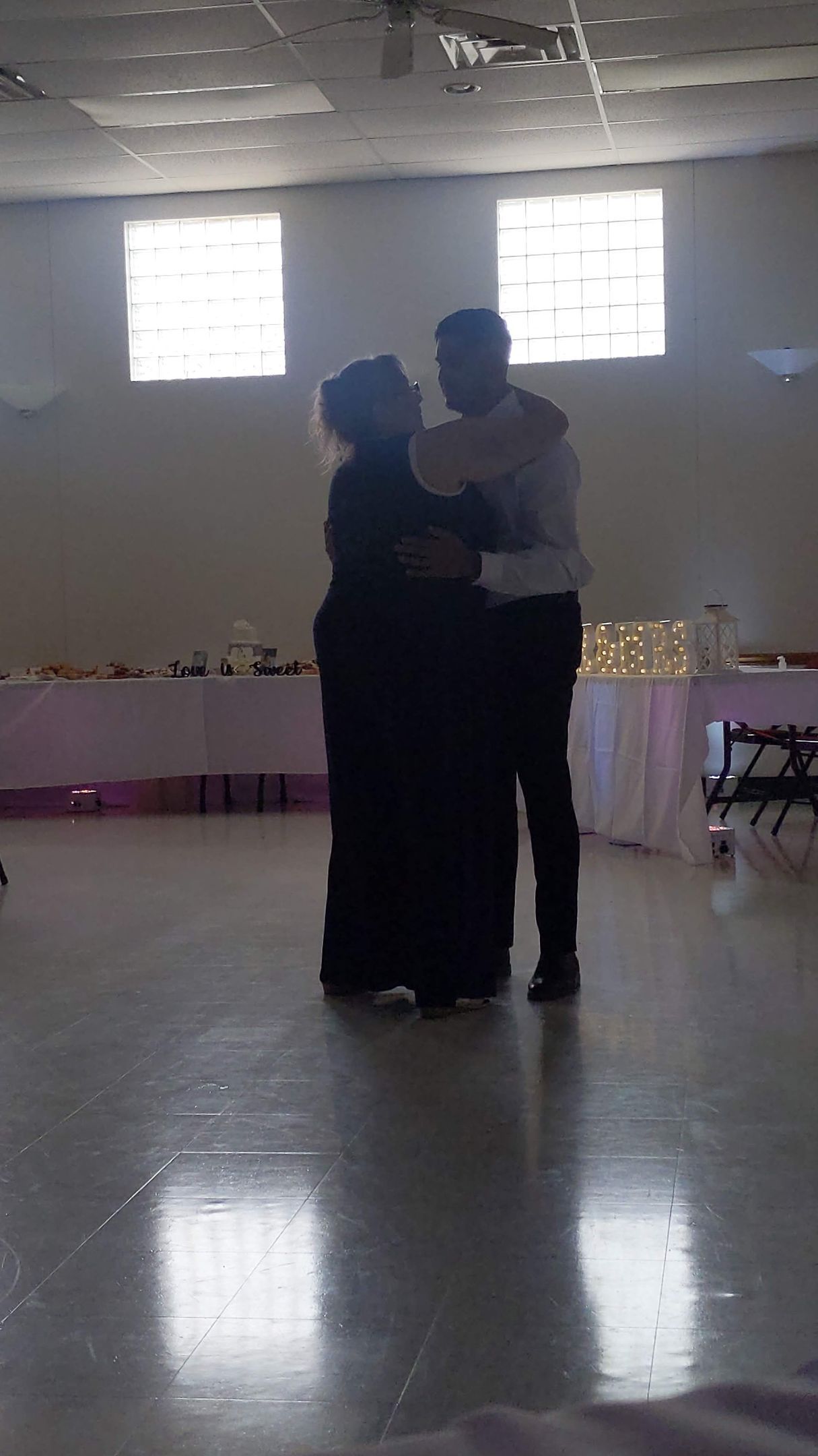 Two people dancing closely in a bright hall with tables and windows in the background.