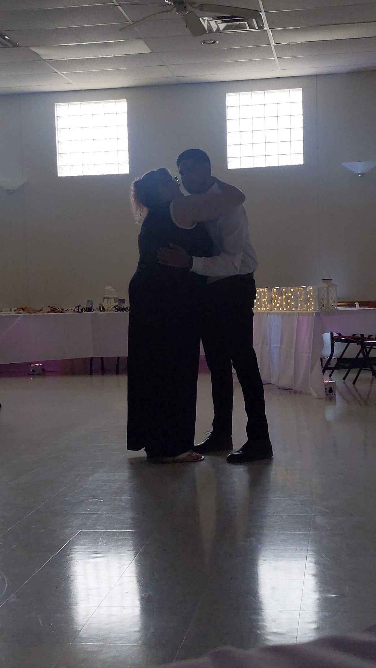 Two people dancing closely in a bright hall with reflective floor and windows behind them